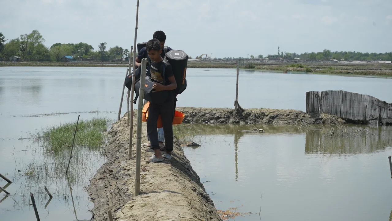 Two young men walking along a narrow, muddy embankment carrying CARE PACKAGE for Emergencies packs.