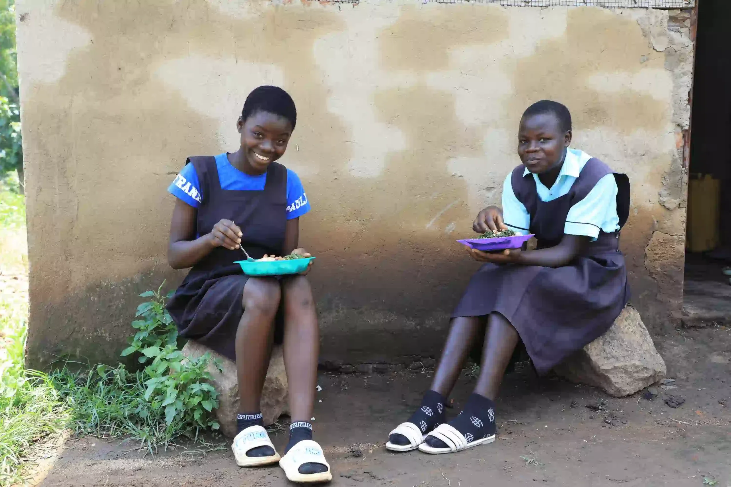 Two smiling students at Ngurwe Primary School in Uganda's Kyangwali Refugee Settlement eating school meals as part of a sustainable school feeding program.