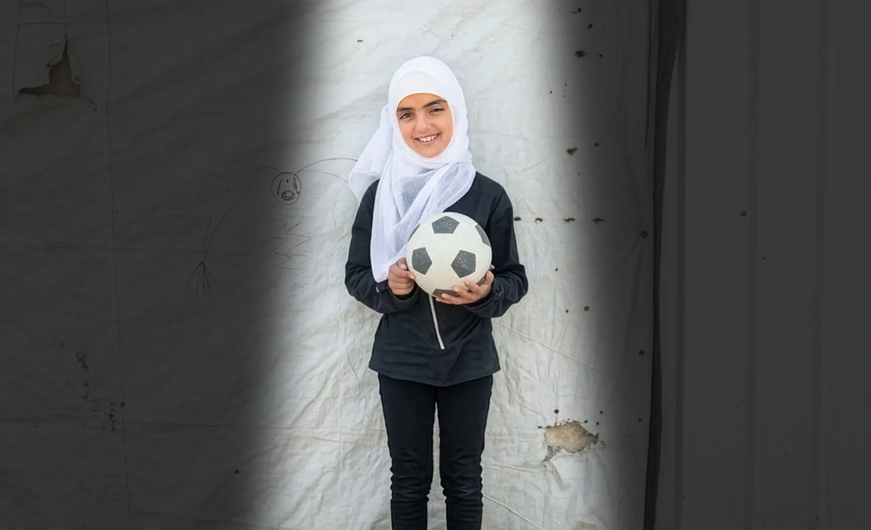 A young girl wearing a white headscarf smiles while holding a soccer ball.
