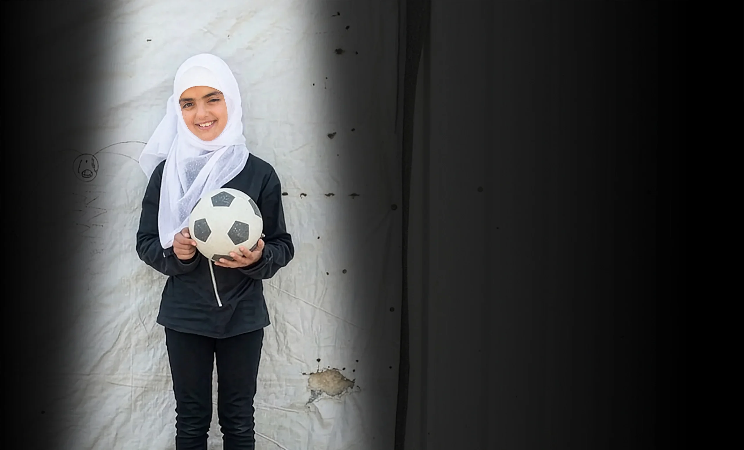 A young girl wearing a white headscarf smiles while holding a soccer ball.