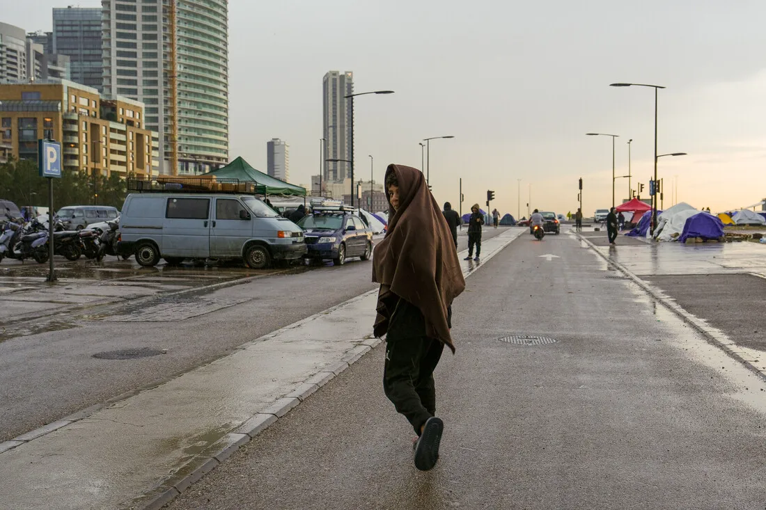 A person wrapped in a brown blanket walks down a wet, empty city street at dusk. Tents and parked vehicles line the sides, with tall buildings nearby.
