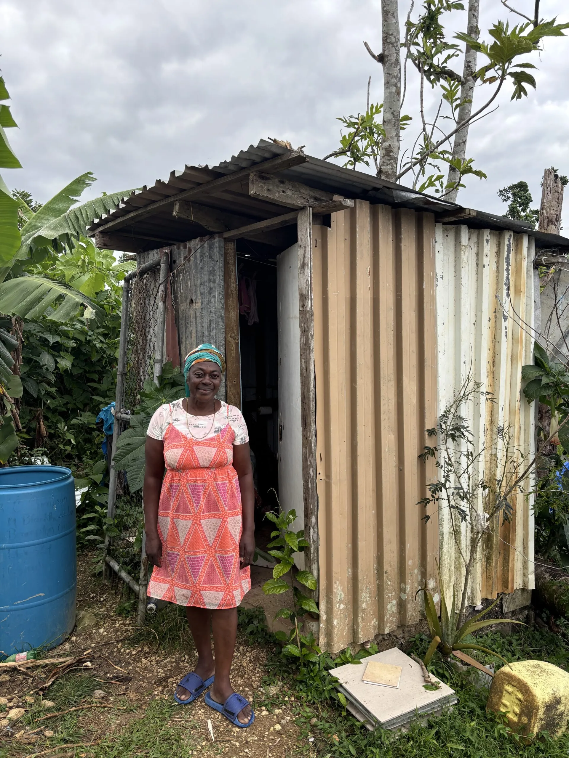 A woman stands next to a small outdoor structure used as shelter during a hurricane, with storm damage visible around her.