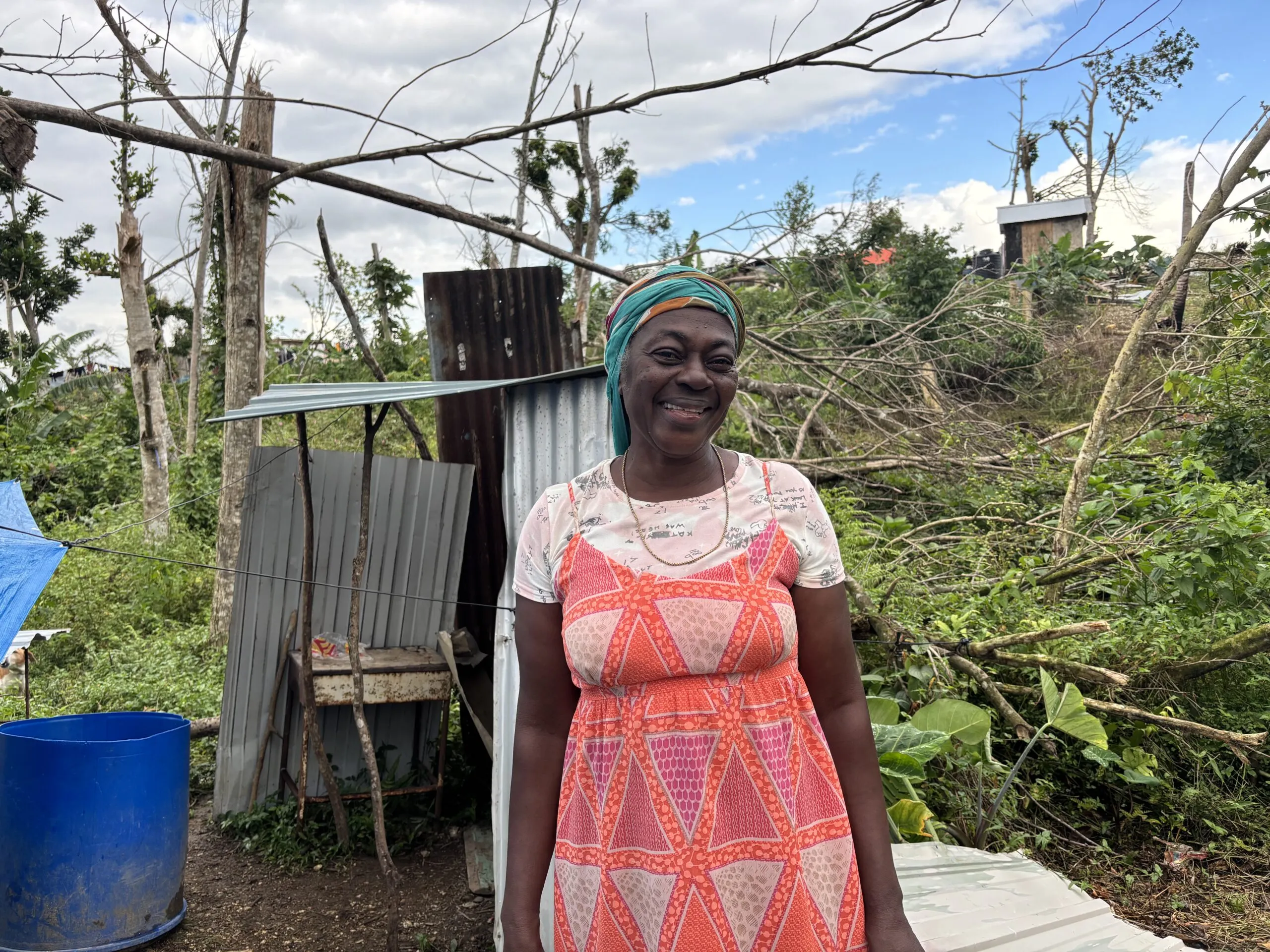 A woman stands outdoors near storm-damaged land and a small structure in rural Jamaica after a hurricane.