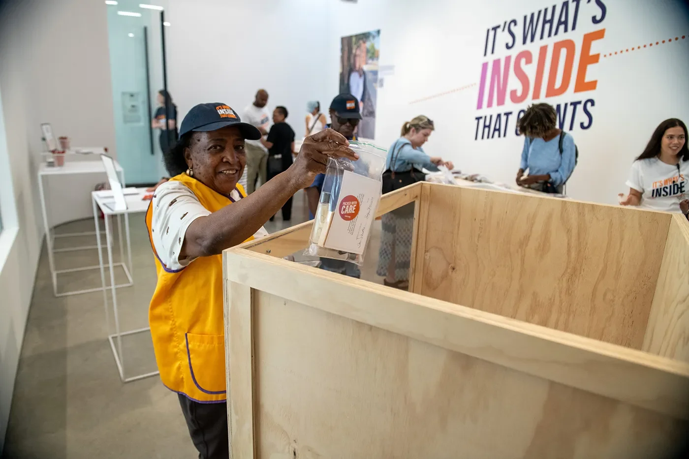 A woman wearing a CARE hat drops a CARE PACKAGE into a large box.