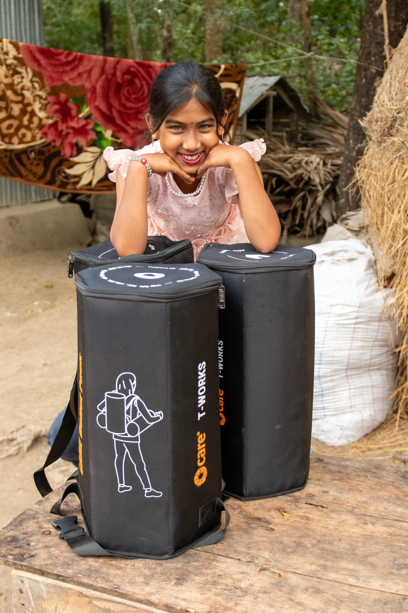 A smiling girl in a pink dress leans on two black containers labeled 