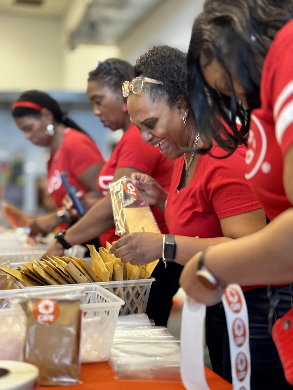 A group of women wearing red t-shirts smile while standing at a table to pack CARE PACKAGE kits.