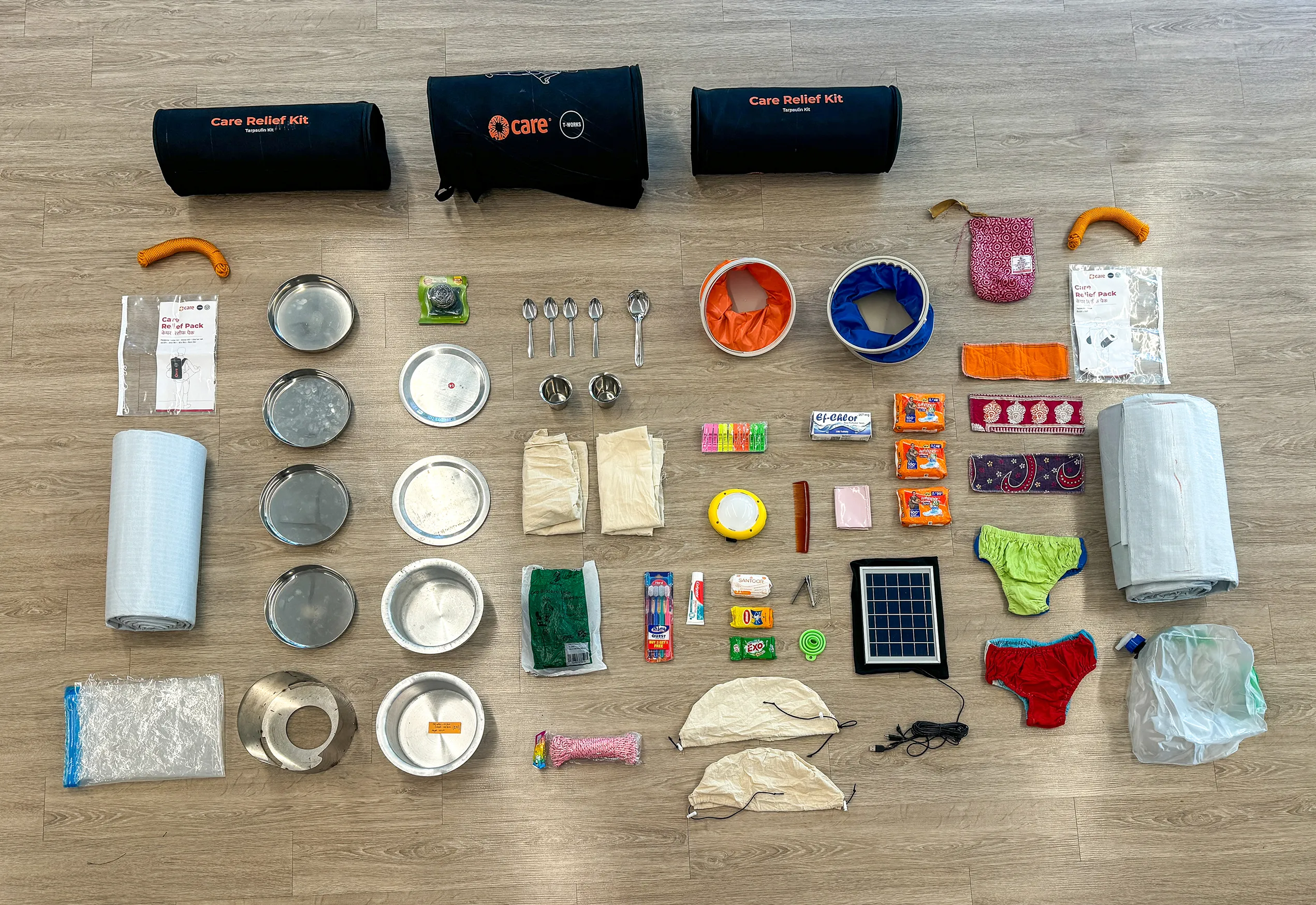 Flat lay showing the contents of a CARE PACKAGE for Emergencies, including shelter materials, cooking supplies, hygiene items, water purification tools, clothing, and solar-powered lighting for a family of four.