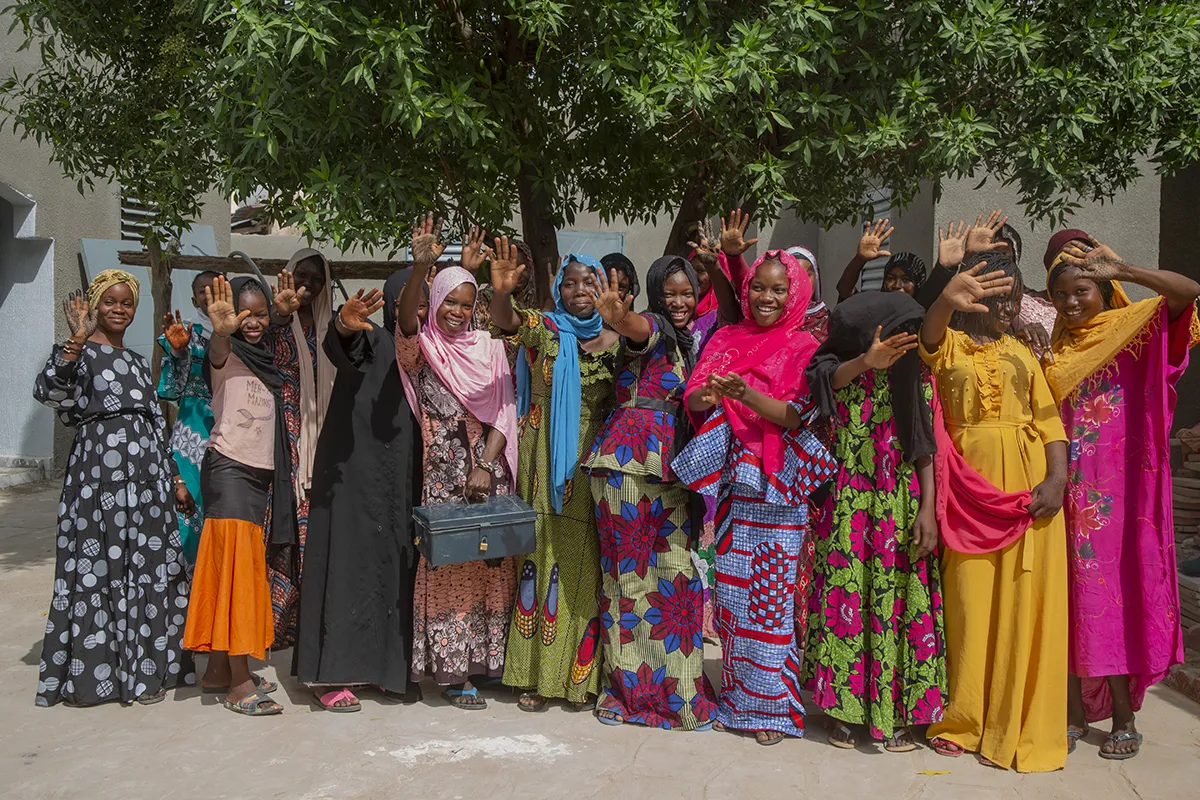 A group of women wearing colorfully patterned clothes smile and wave.