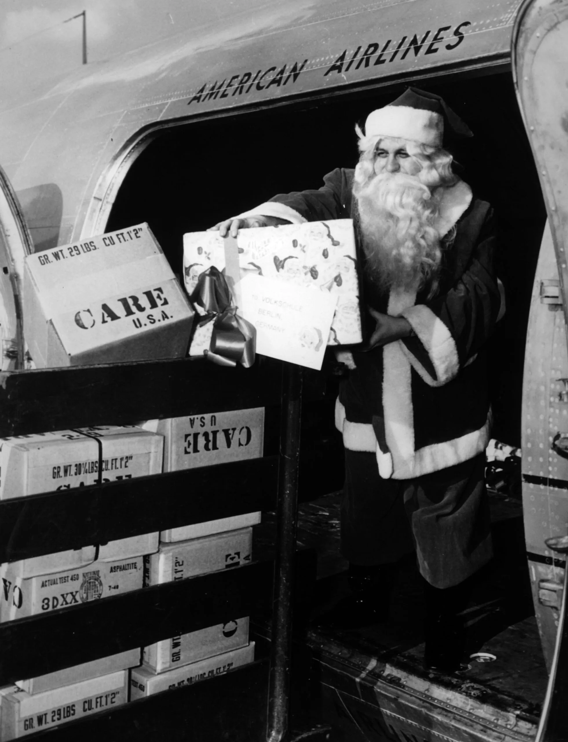 A black and white photo of a man dressed like Santa Claus unloading CARE PACKAGE boxes from an airplane.