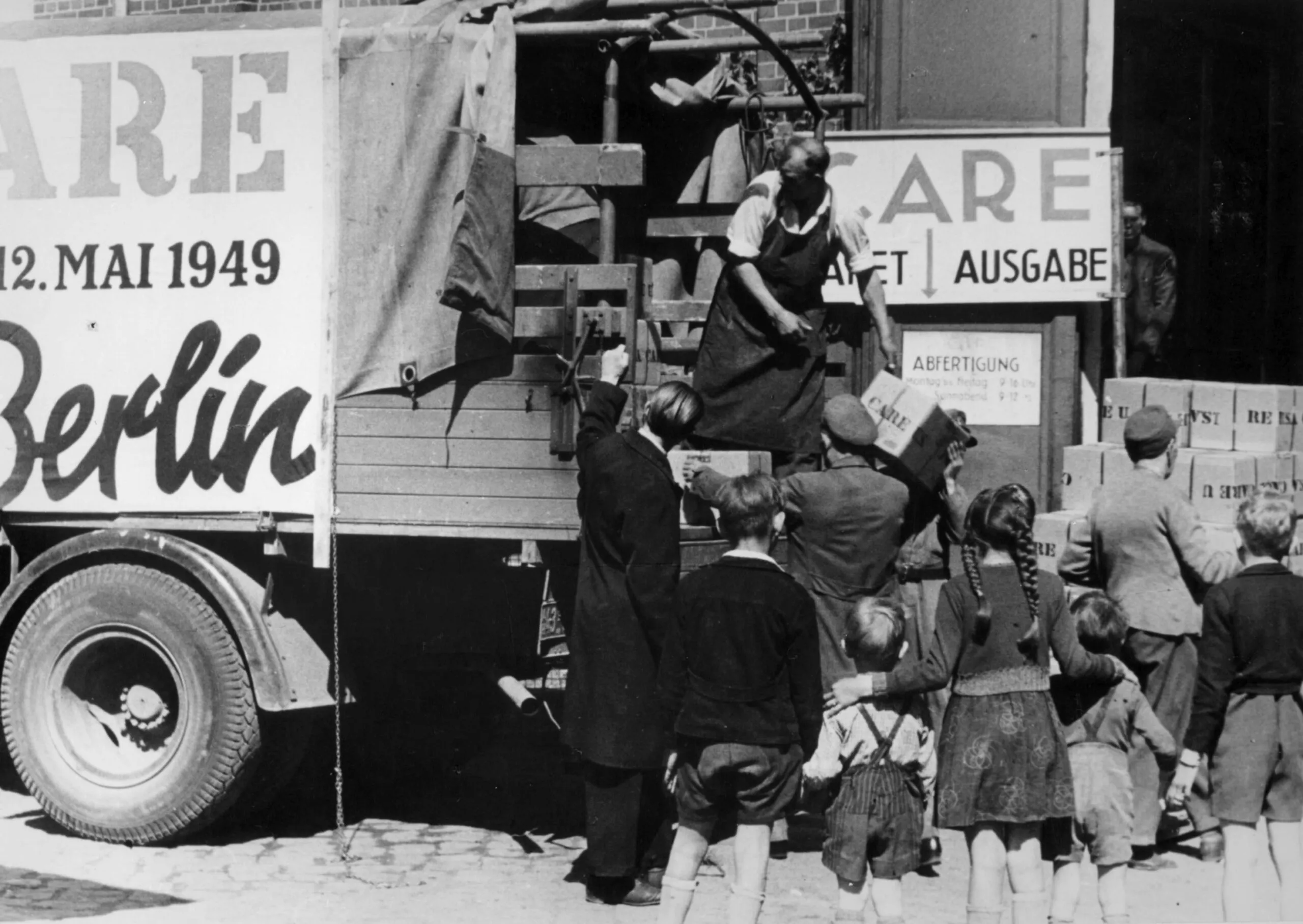 A group of people gather around a truck as a man unloads CARE PACKAGE parcels from the back.