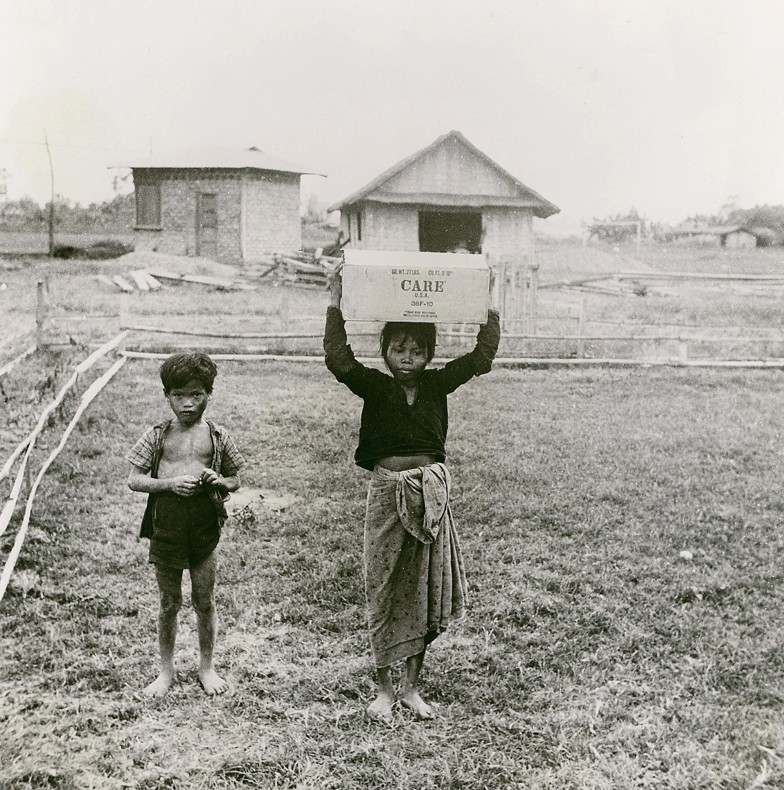 A young boy and girl walking through a rural area in the Philippines. The girl is carrying a CARE PACKAGE on top of her head.