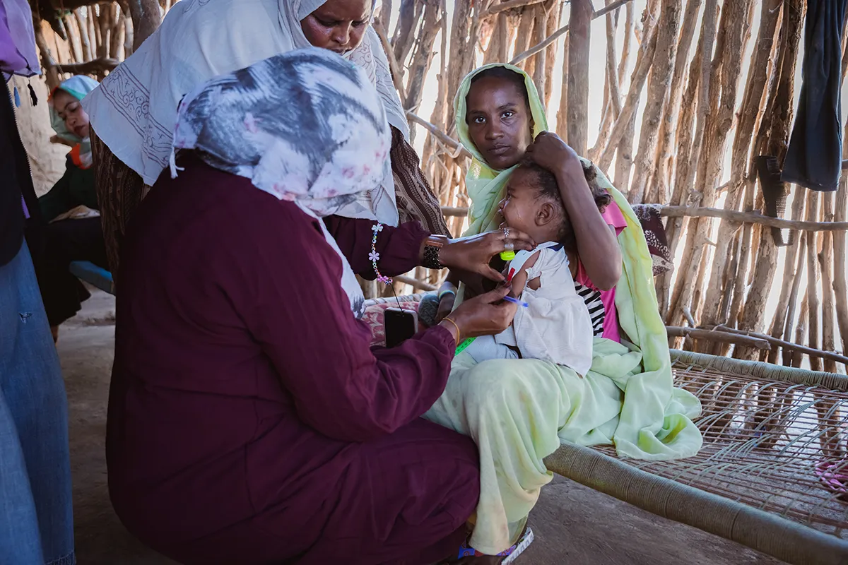 A woman holds onto a crying young child while a female healthcare worker measures the child's arm width.