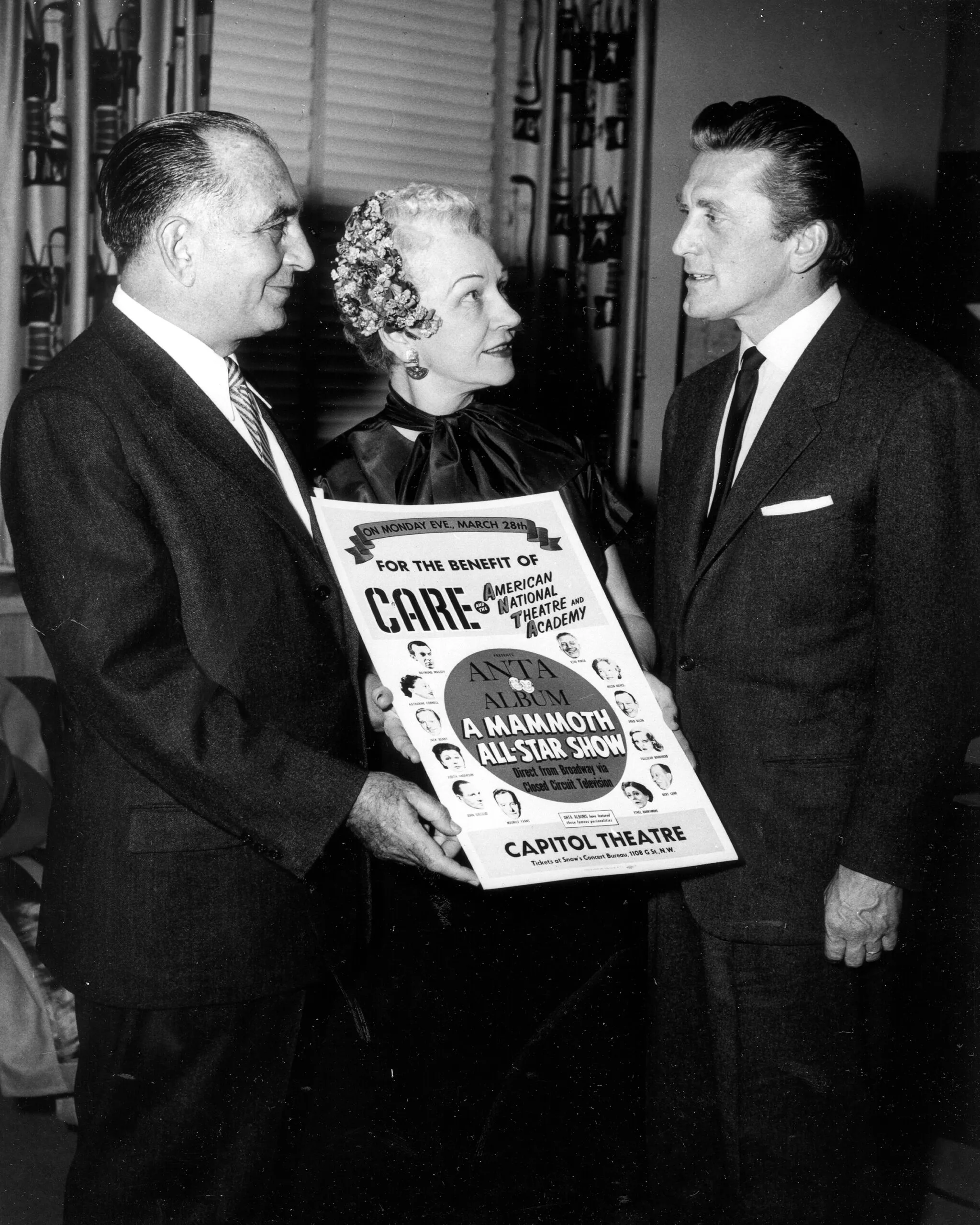 Actor Kirk Douglas standing next to a man and woman who are holding a sign for a CARE benefit show.