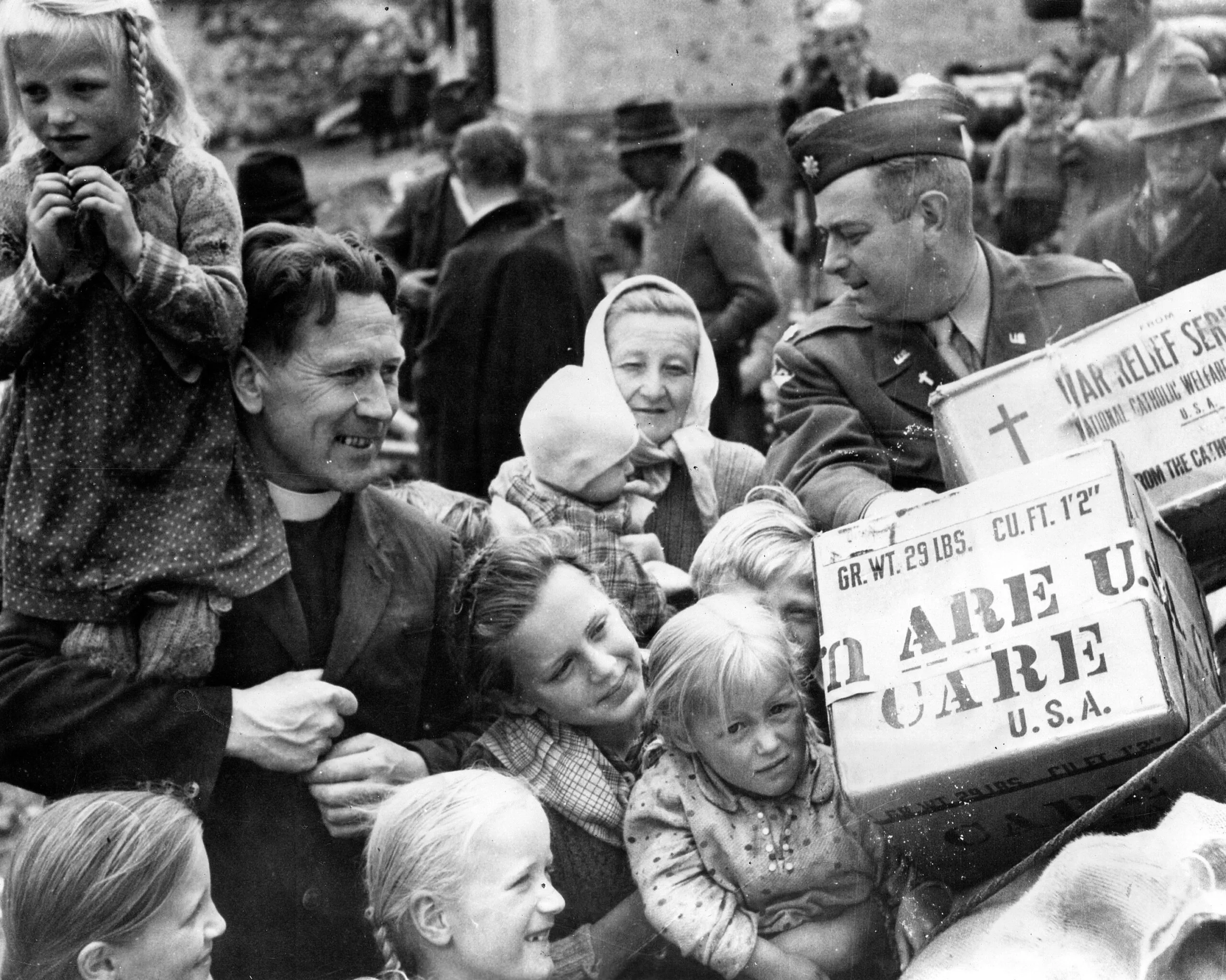 A black and white photo of a Reverend with a group of children and women being handed a CARE PACKAGE by a man in a military uniform.