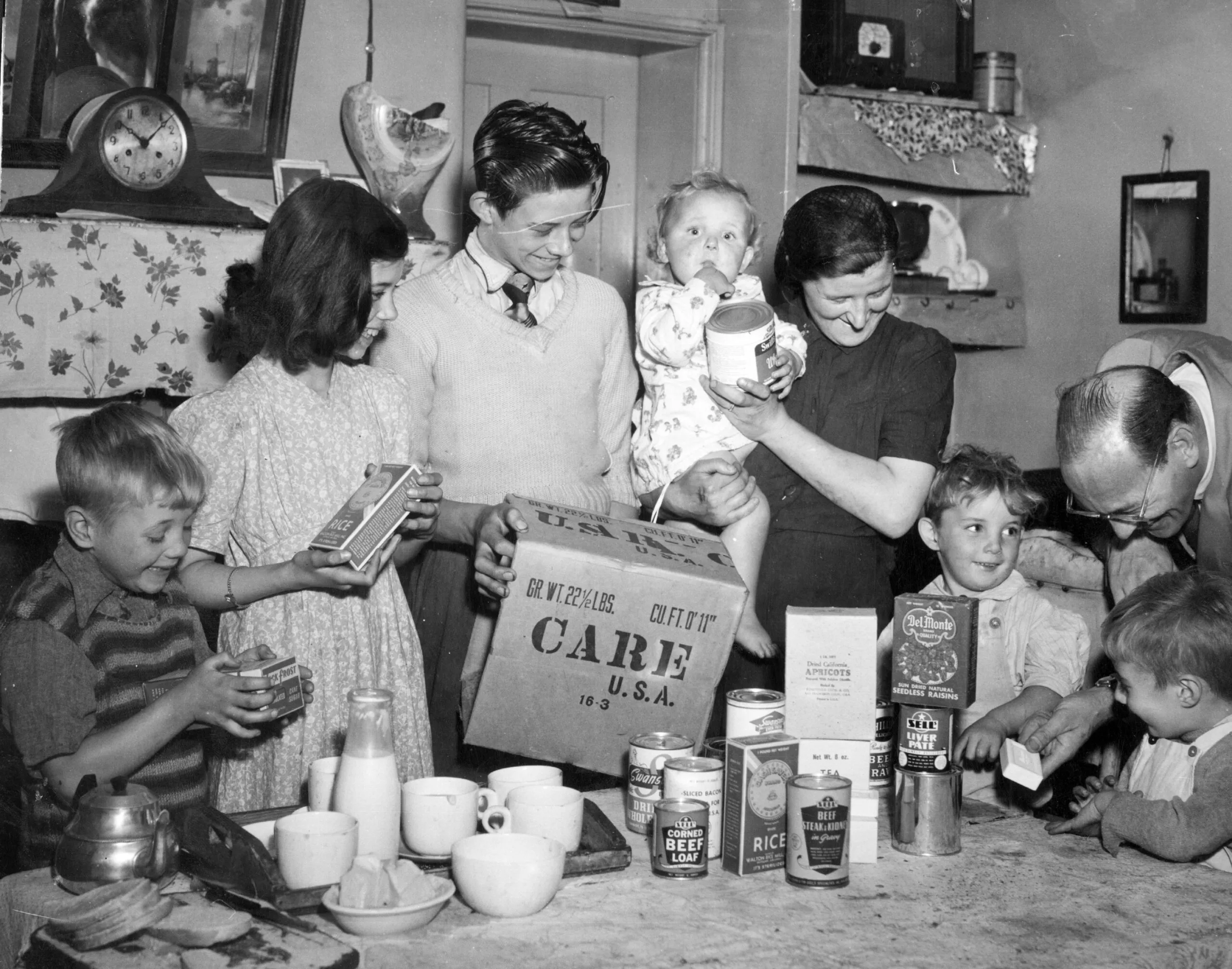 A black and white photo of a mother and her six children in their kitchen looking at the items from a CARE PACKAGE that a CARE worker has given them.