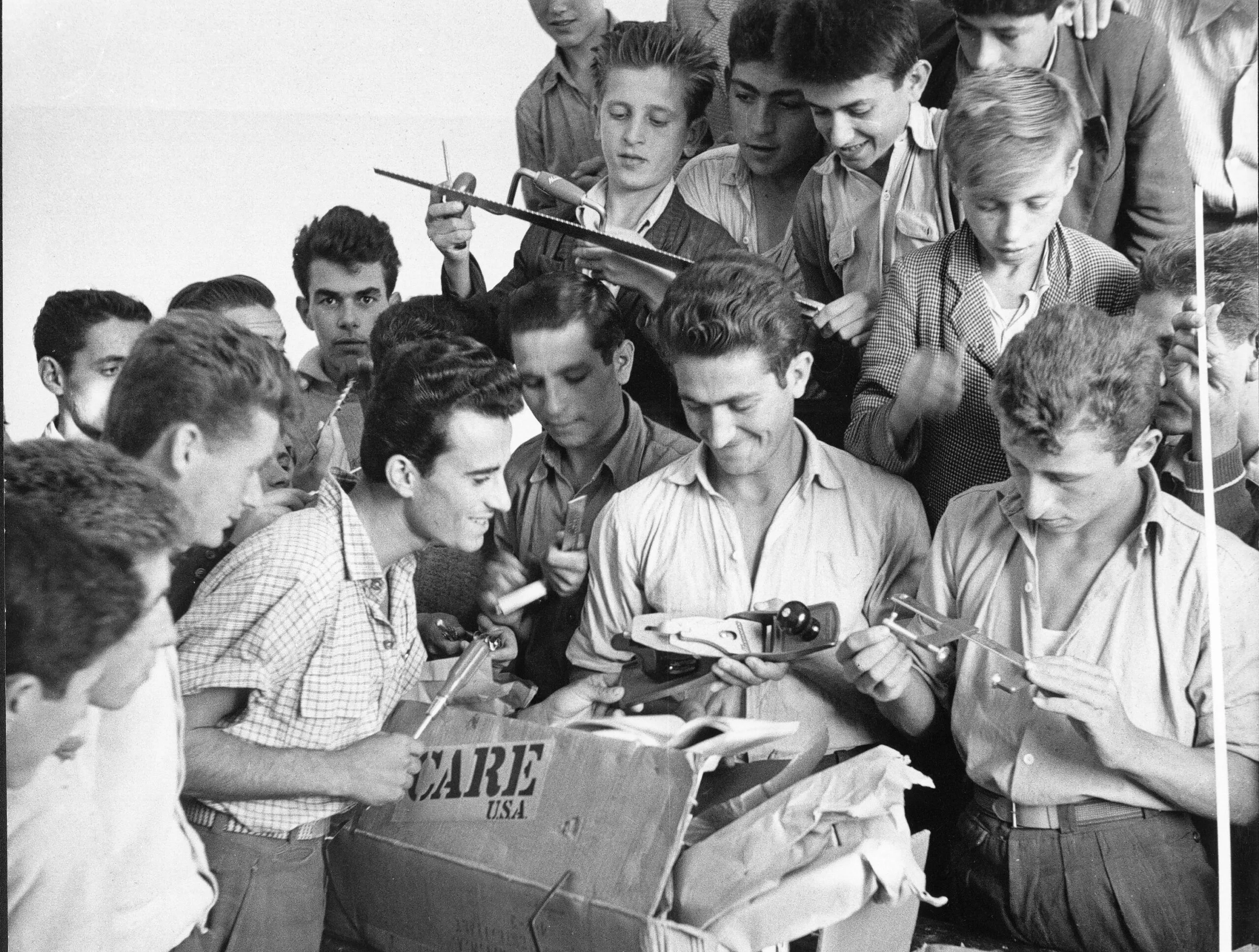 A black and white photo of a group of young men opening a CARE PACKAGE and looking at the new tools.