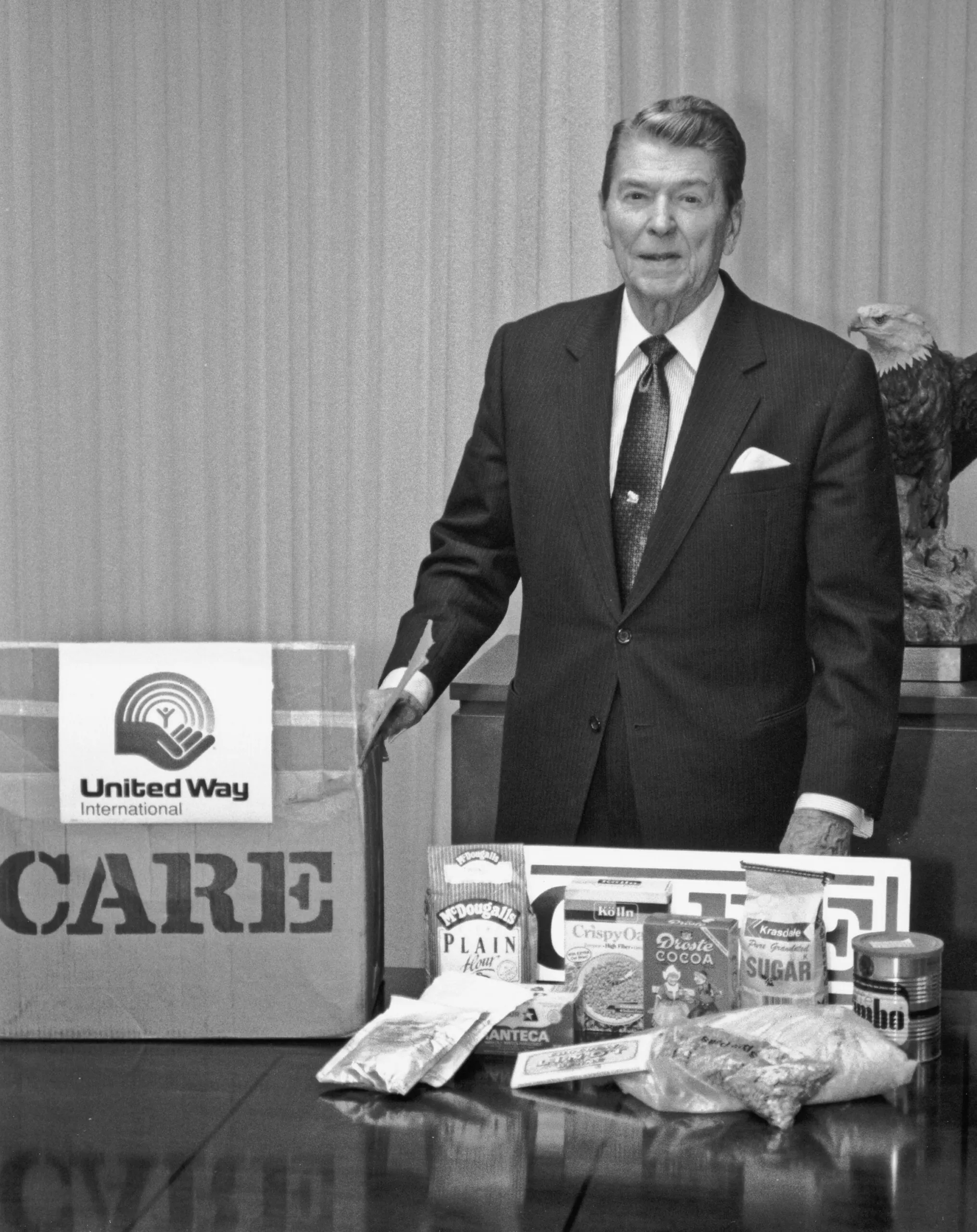 President Ronald Reagan standing in front of a CARE PACKAGE box with food items laid out on a table.