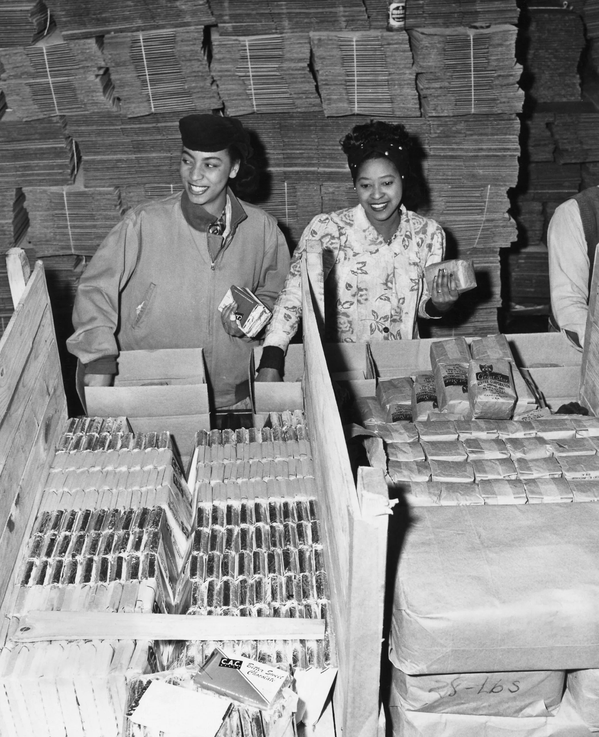 A black and white photo of two women in a warehouse putting items inside CARE PACKAGE boxes.