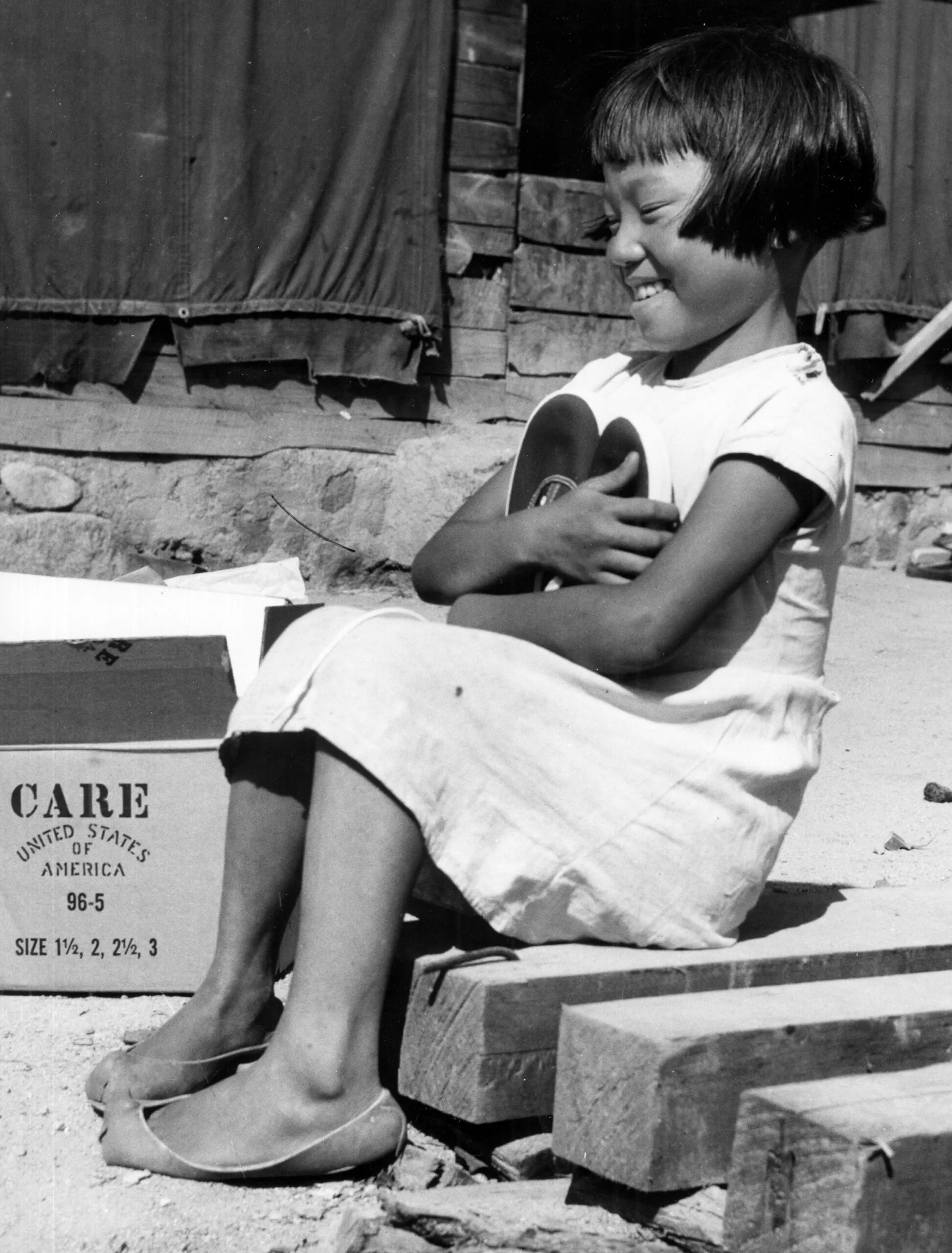 A black and white photo of a young girl sitting on a wooden block, smiling and hugging a pair of new shoes from a CARE PACKAGE.