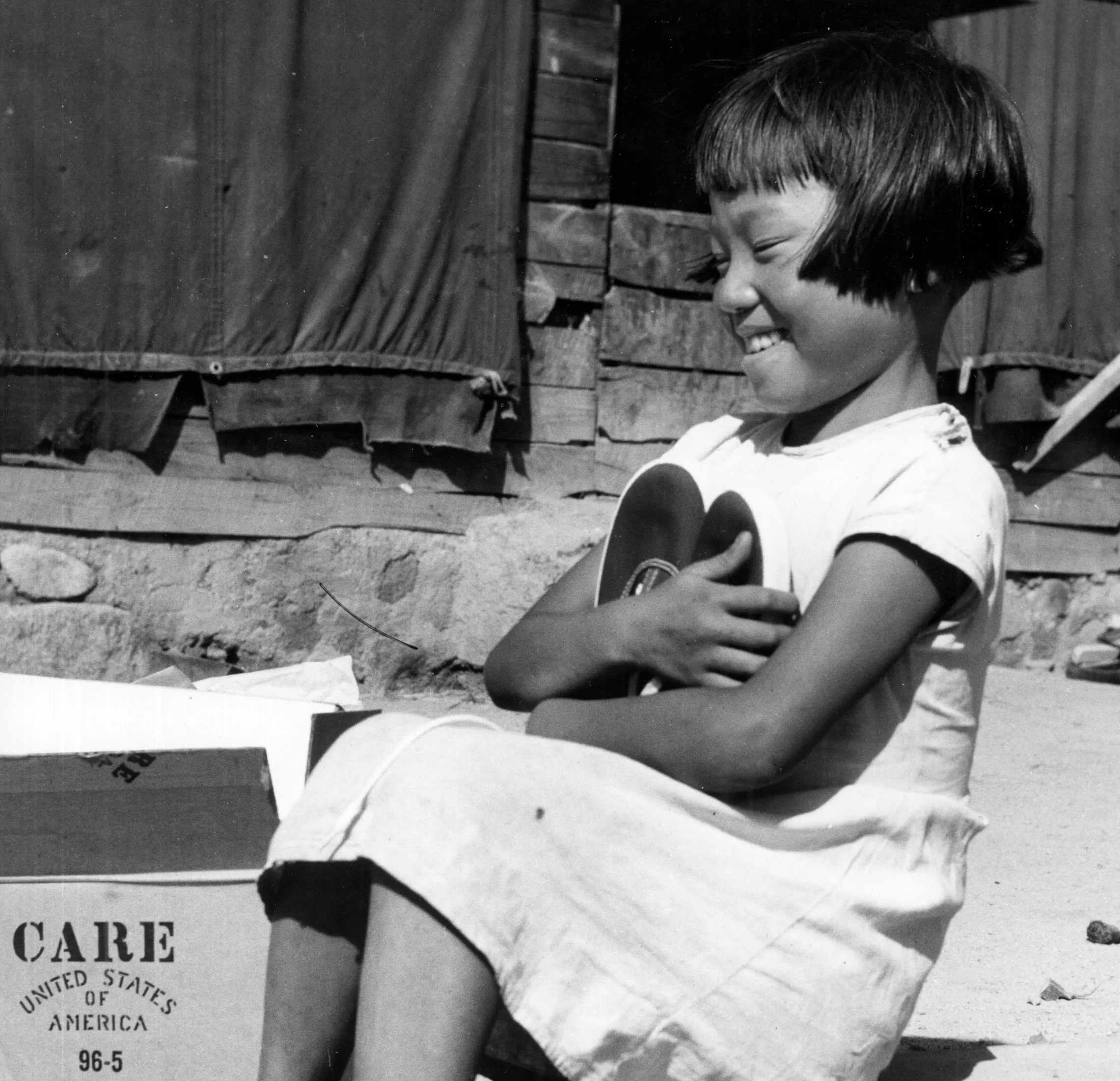 A young Korean girl sits on a curb and smiles while hugging a pair of shoes.