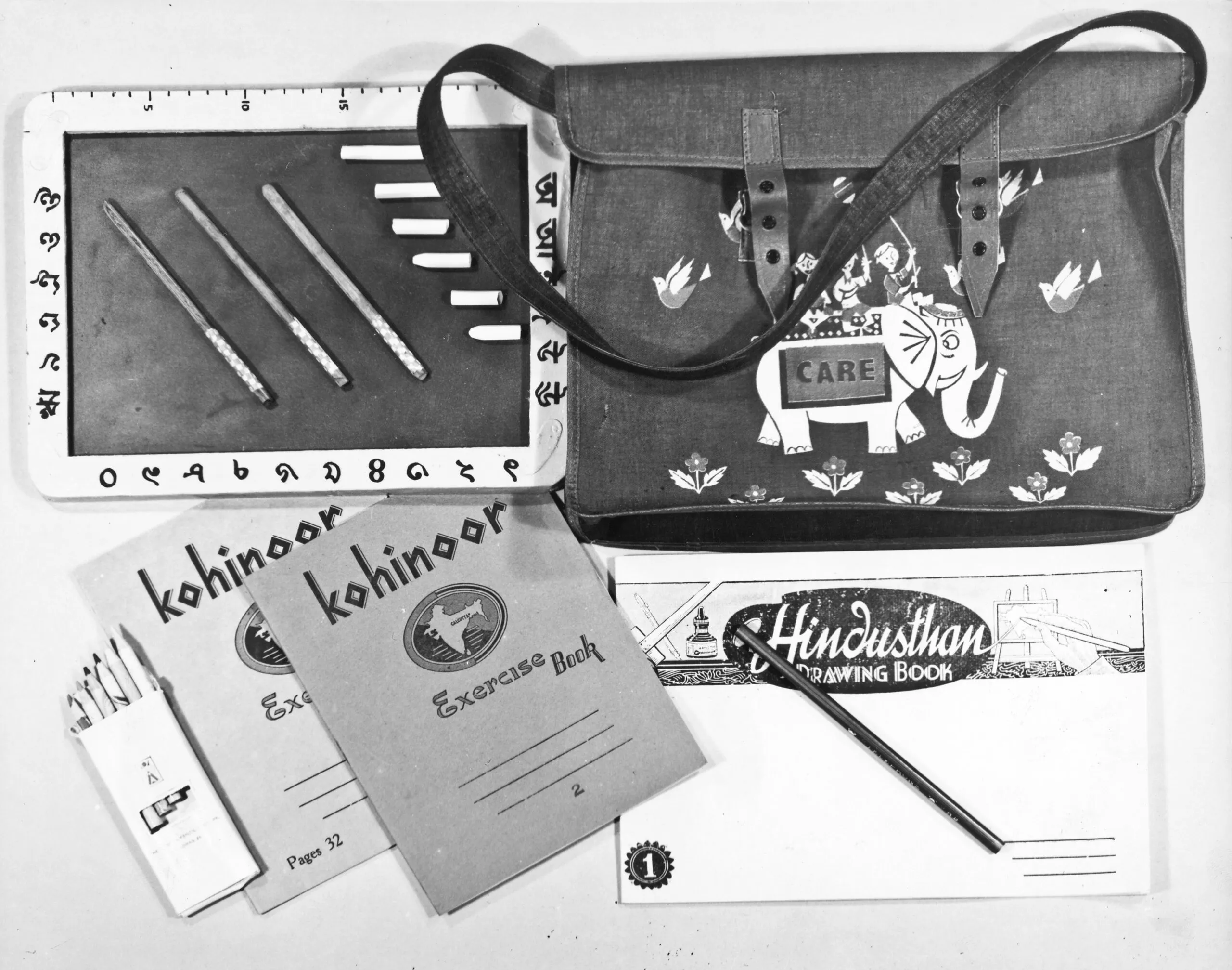 A black and white photo of items from a CARE PACKAGE school kit. It contains pencils, a school bag, a drawing book and writing books.