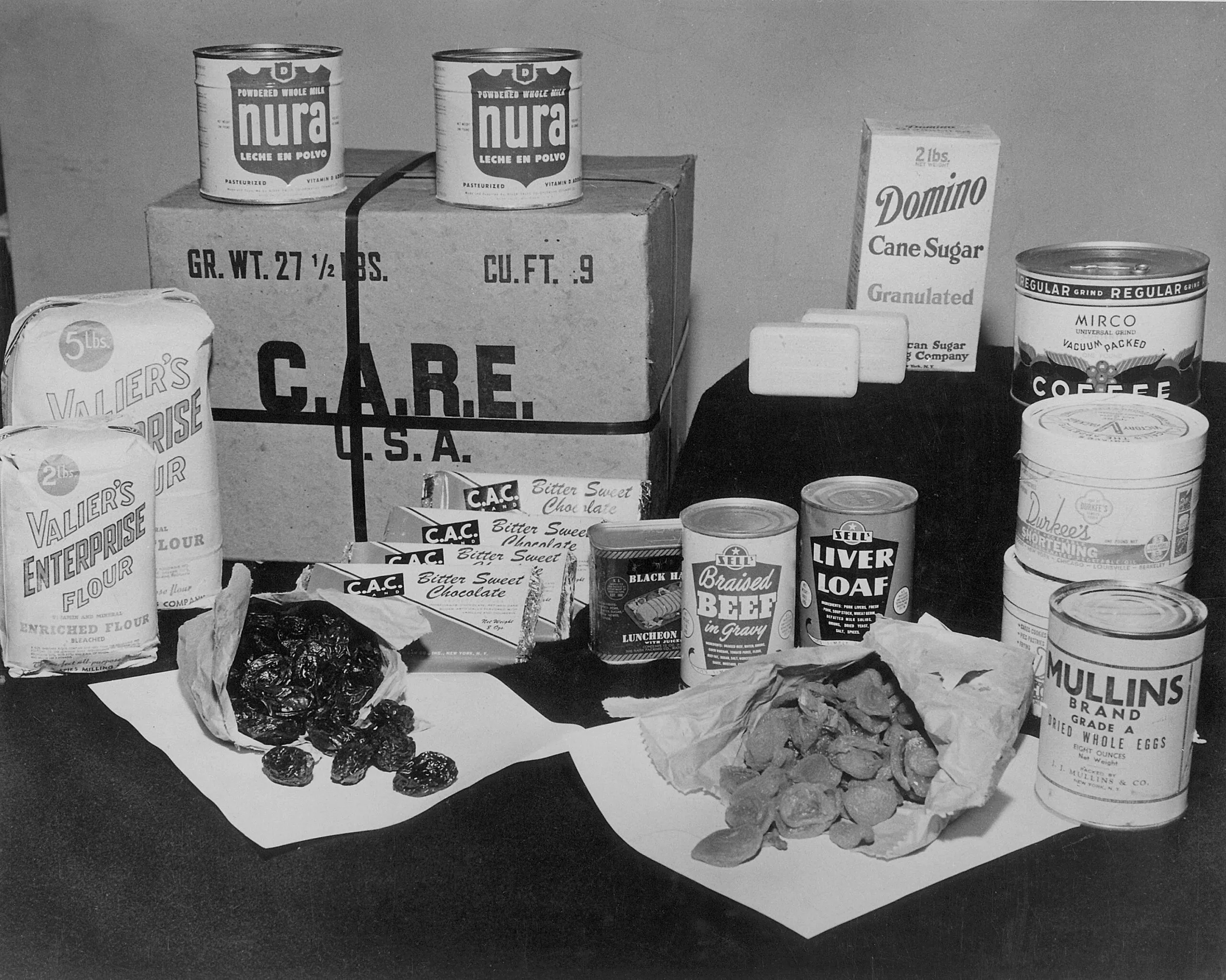 A historical image of items found in a CARE PACKAGE from the 1940s. The items are positioned on a table and include: cans of meat, bags of sugar, bars of chocolate, a can of coffee and dried fruit.