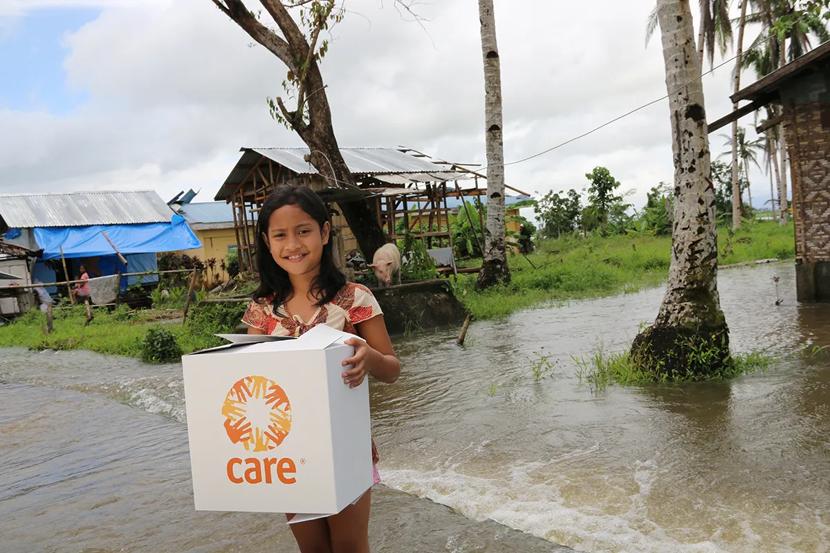 A young girl smiles while holding a CARE PACKAGE near a flooded road.