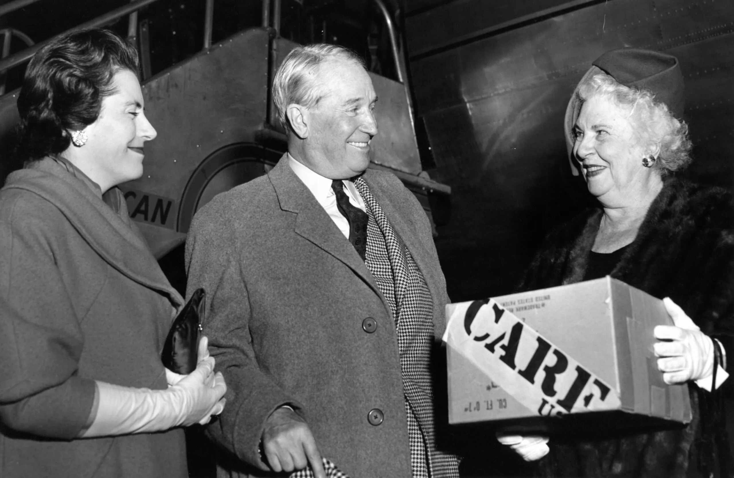 French actor Maurice Chevalier stands next to two women. The woman on the right is holding a CARE PACKAGE.