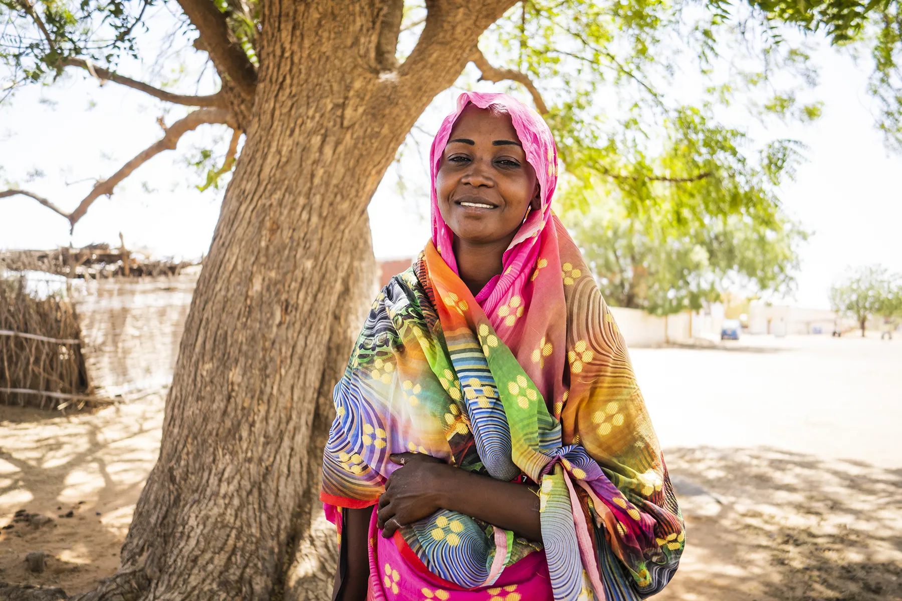 A woman wearing a multi-colored cloth smiles while standing next to a tree.