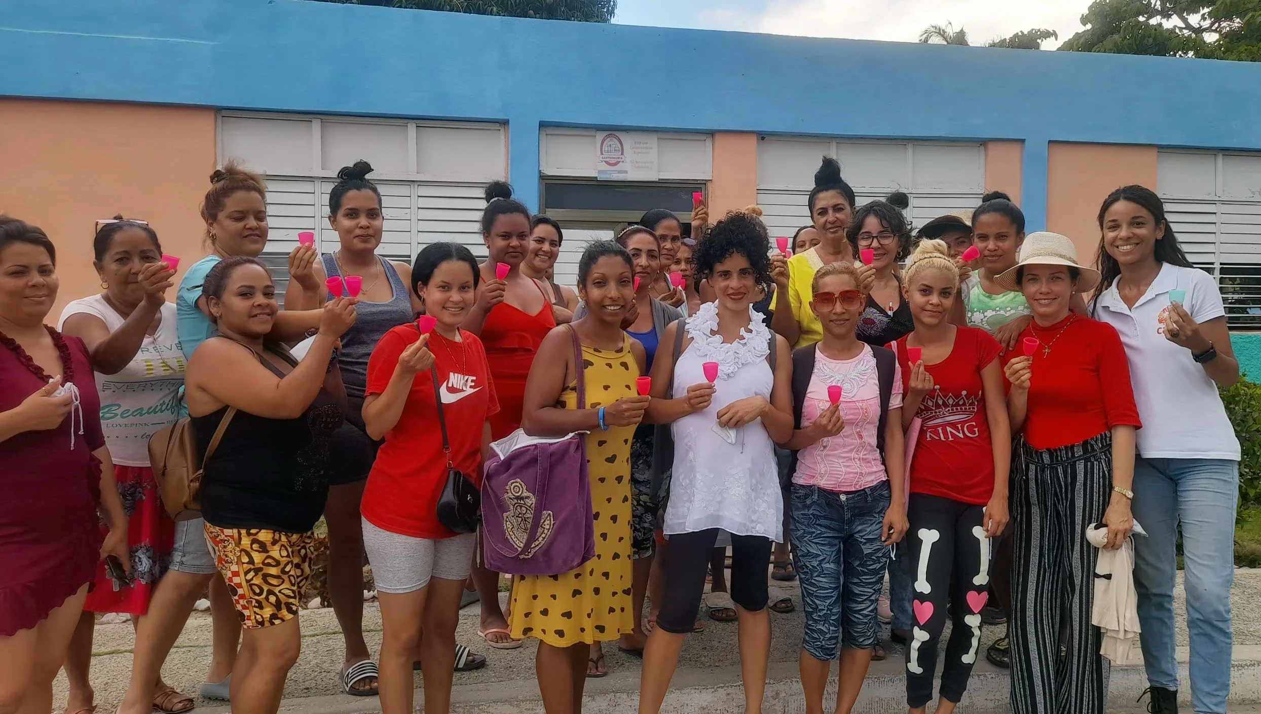 A group of women in Imías, Guantánamo, Cuba, standing together outdoors and smiling while holding up small, pink menstrual cups as part of a community health workshop.