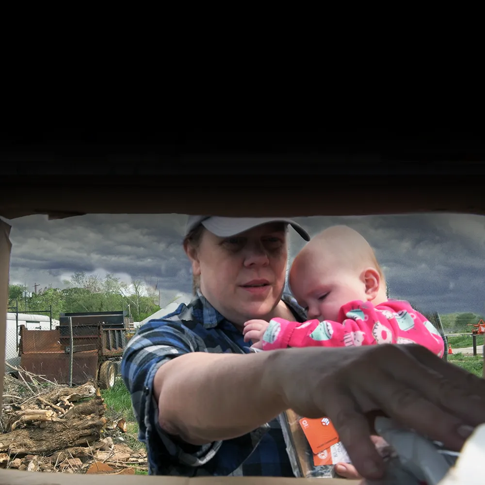 A woman holding a small baby reaches into a box.
