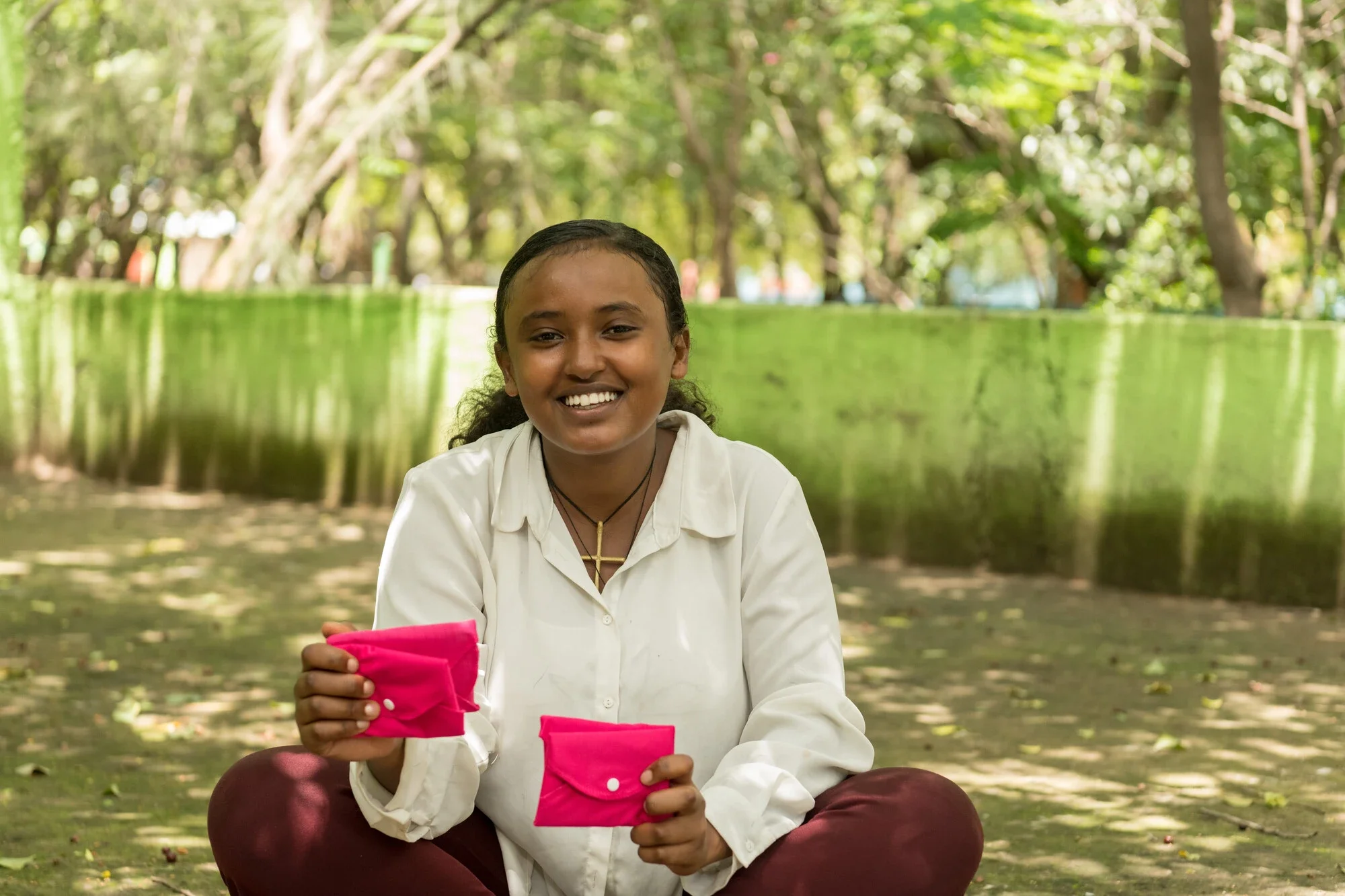 A smiling young Ethiopian woman named Sara sitting outdoors on the grass, holding two pink reusable sanitary pads towards the camera.