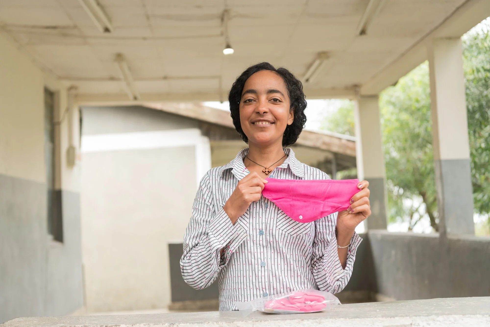 A smiling young woman in Ethiopia holding up a reusable pink sanitary pad, with a packaged set resting on the surface in front of her.