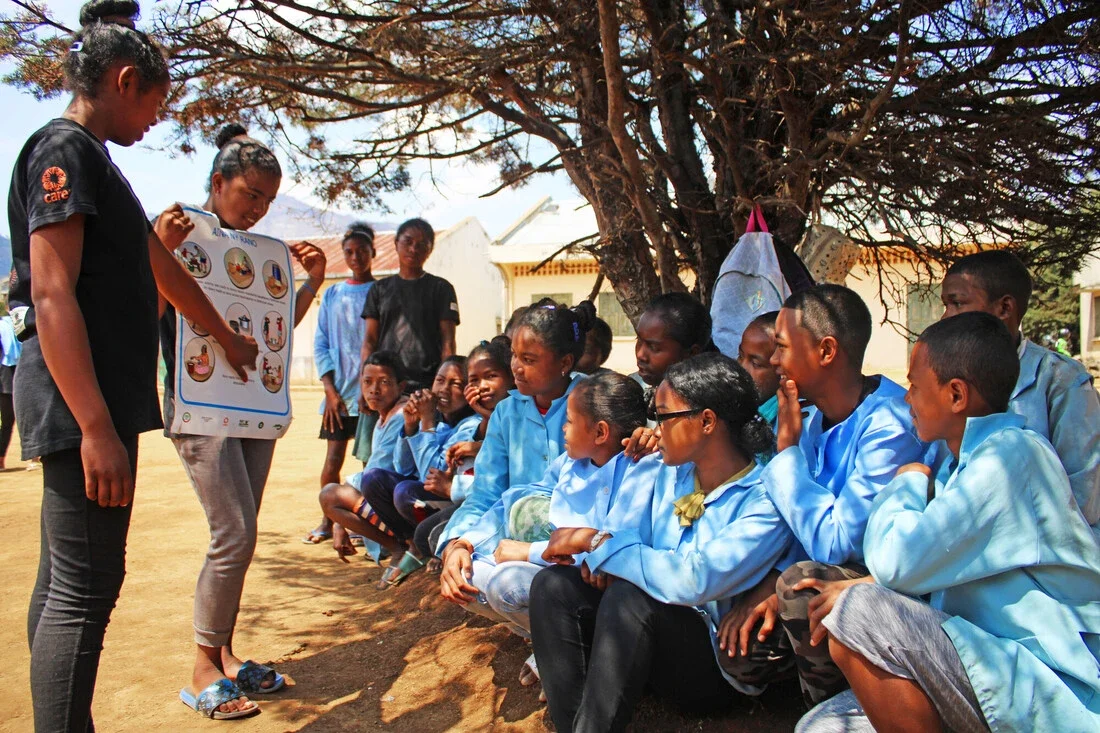 Under the shade of a tree, two young women — one wearing a 'CARE' logo T-shirt — lead a menstrual health discussion using an illustrated chart for a group of attentive schoolgirls and boys in blue uniforms seated on the ground.