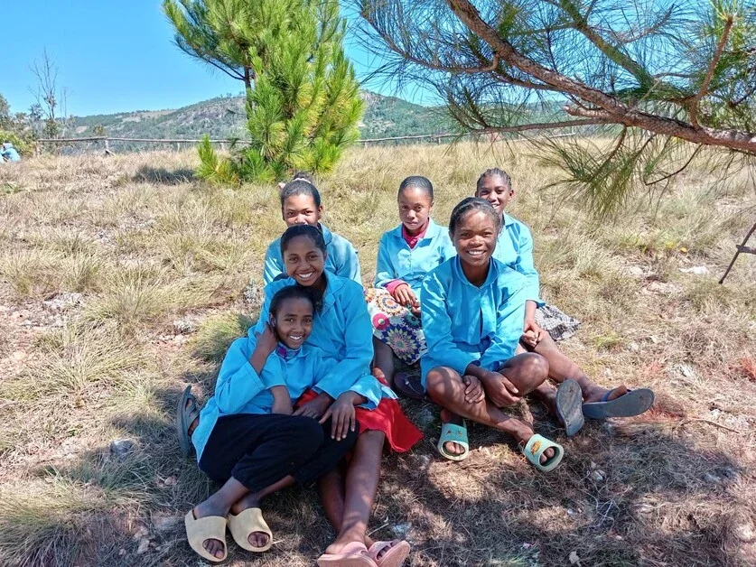 Five Ethiopian schoolgirls in blue uniforms sitting together outdoors under a tree, smiling.