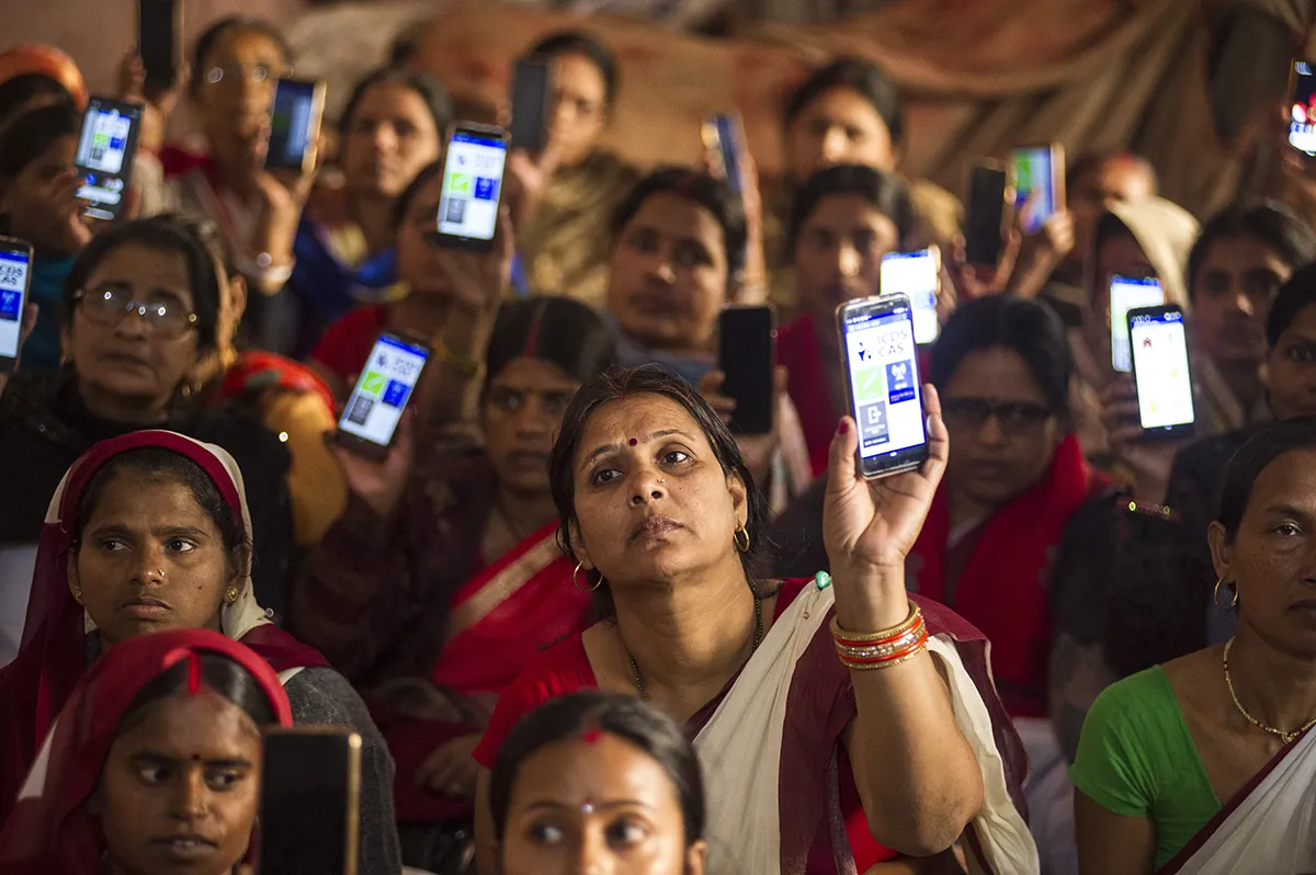 A large group of women in Bihar, India, hold up their mobile phones open to a health app.