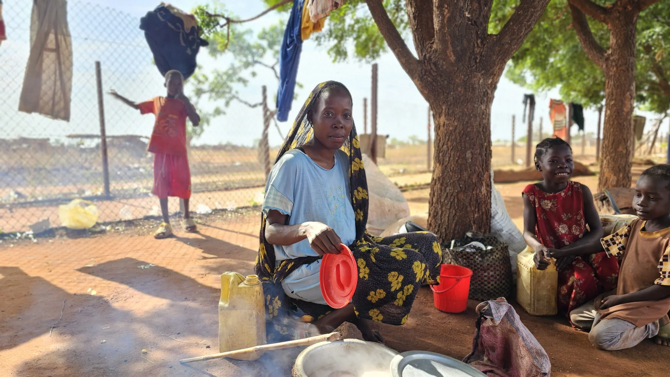 A woman prepares rice over a fire while her children rest in the shade.