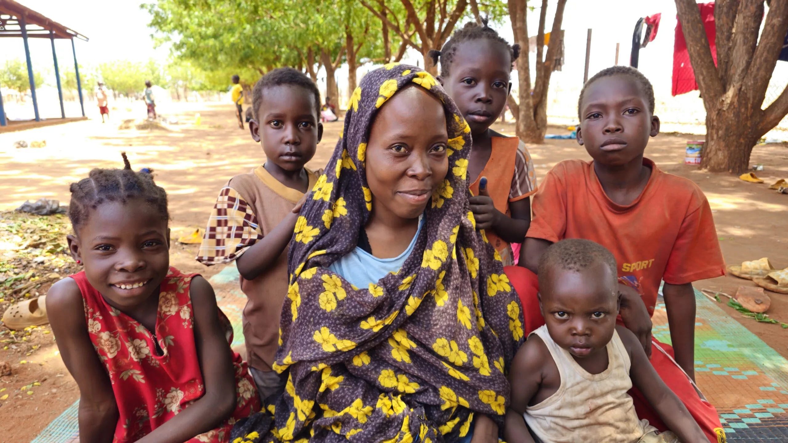 A family — a mother and five children — look at the camera as they sit on a colorful woven mat at a refugee reception center in Yida, South Sudan.