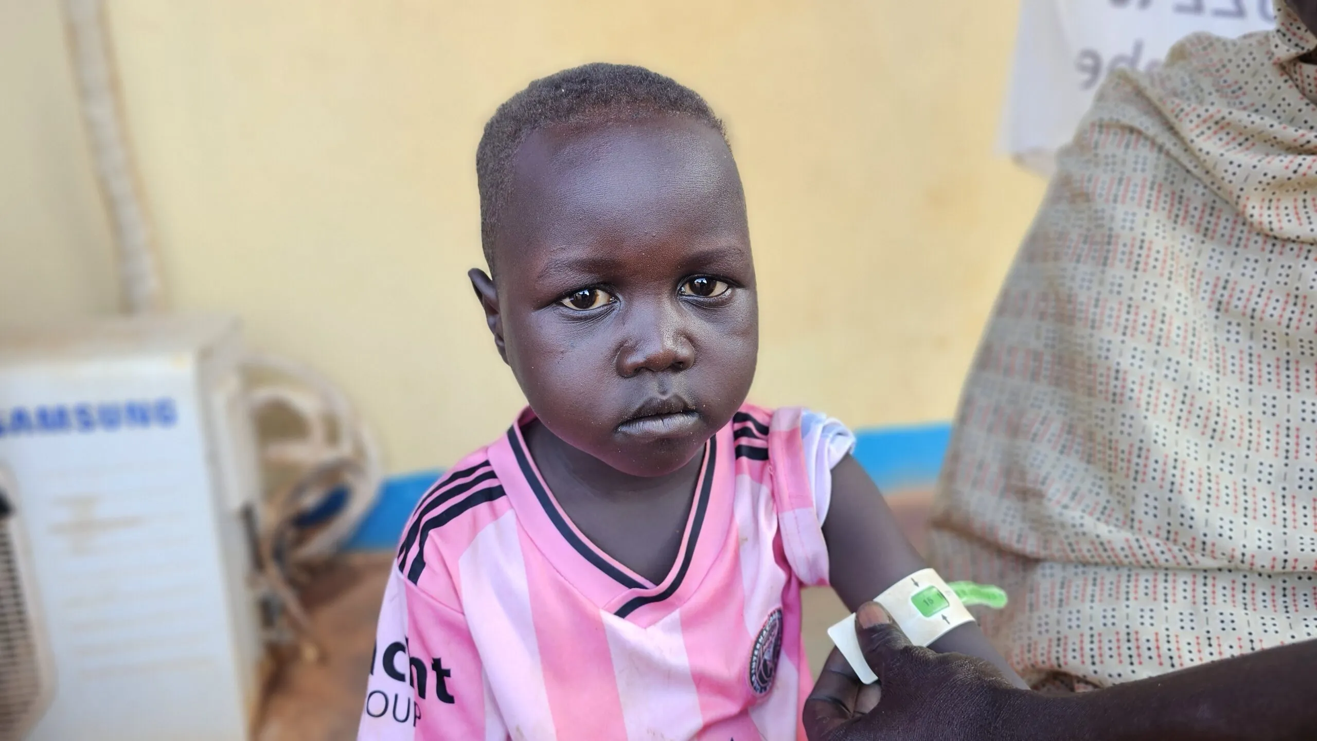 A boy in a striped, pink soccer jersey looks into the camera. Around his upper arm, a MUAC tape shows that he is recovering well after treatment for severe malnutrition.