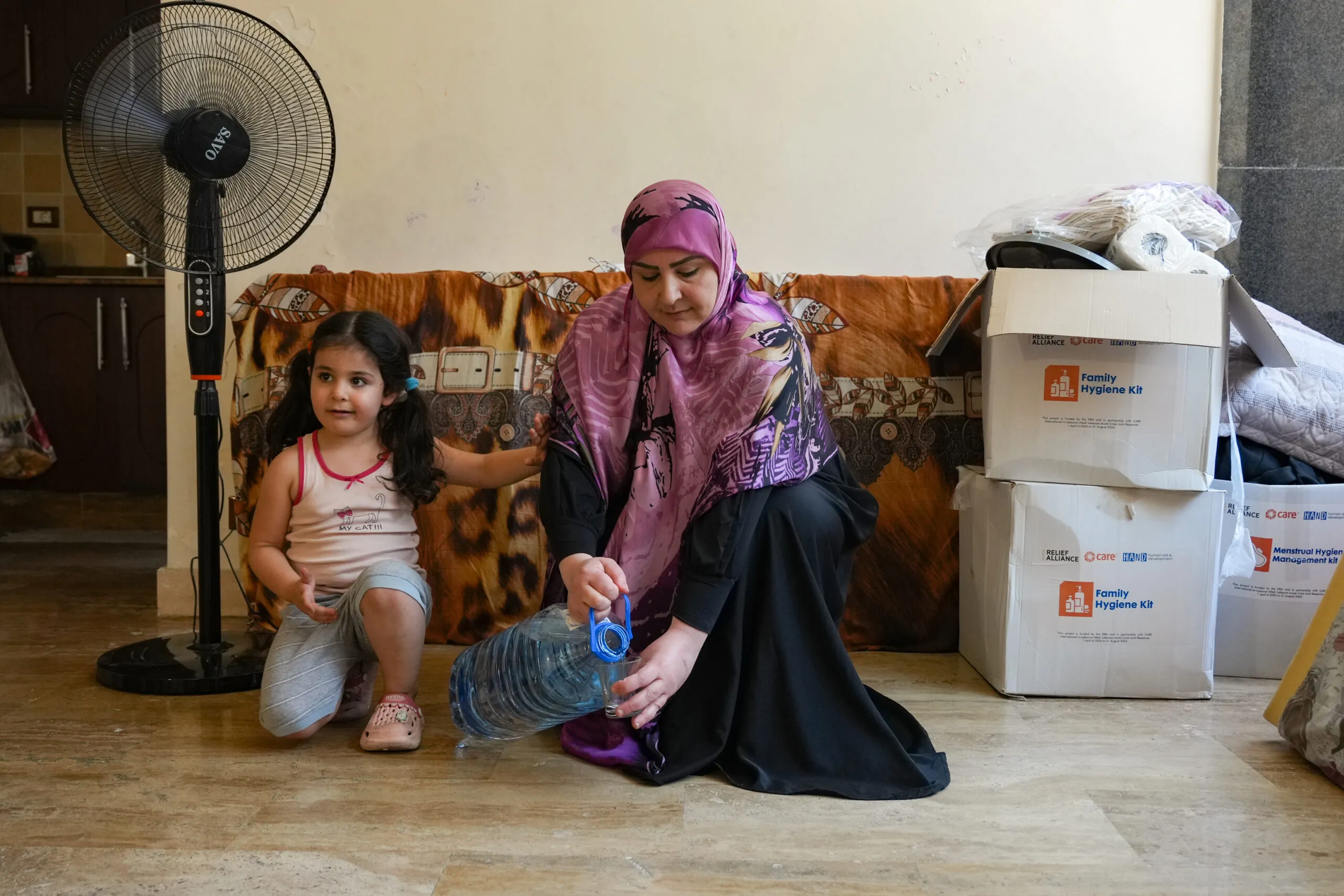 A woman is pouring a glass of water as her young daughter sits on the floor next to her inside an apartment.