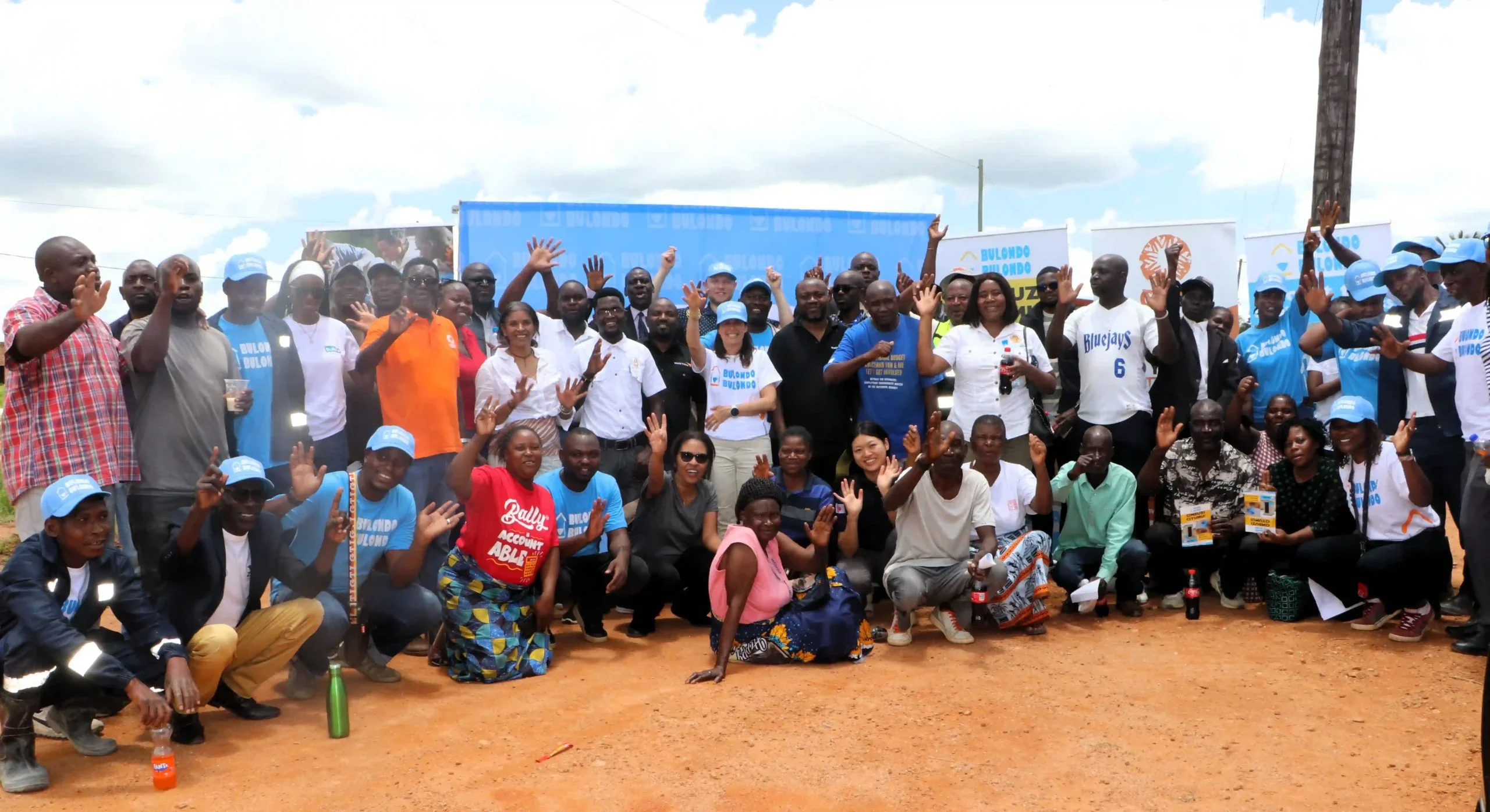 A large, diverse group of people smiling and waving at the camera during an outdoor community event. Many individuals are wearing blue 