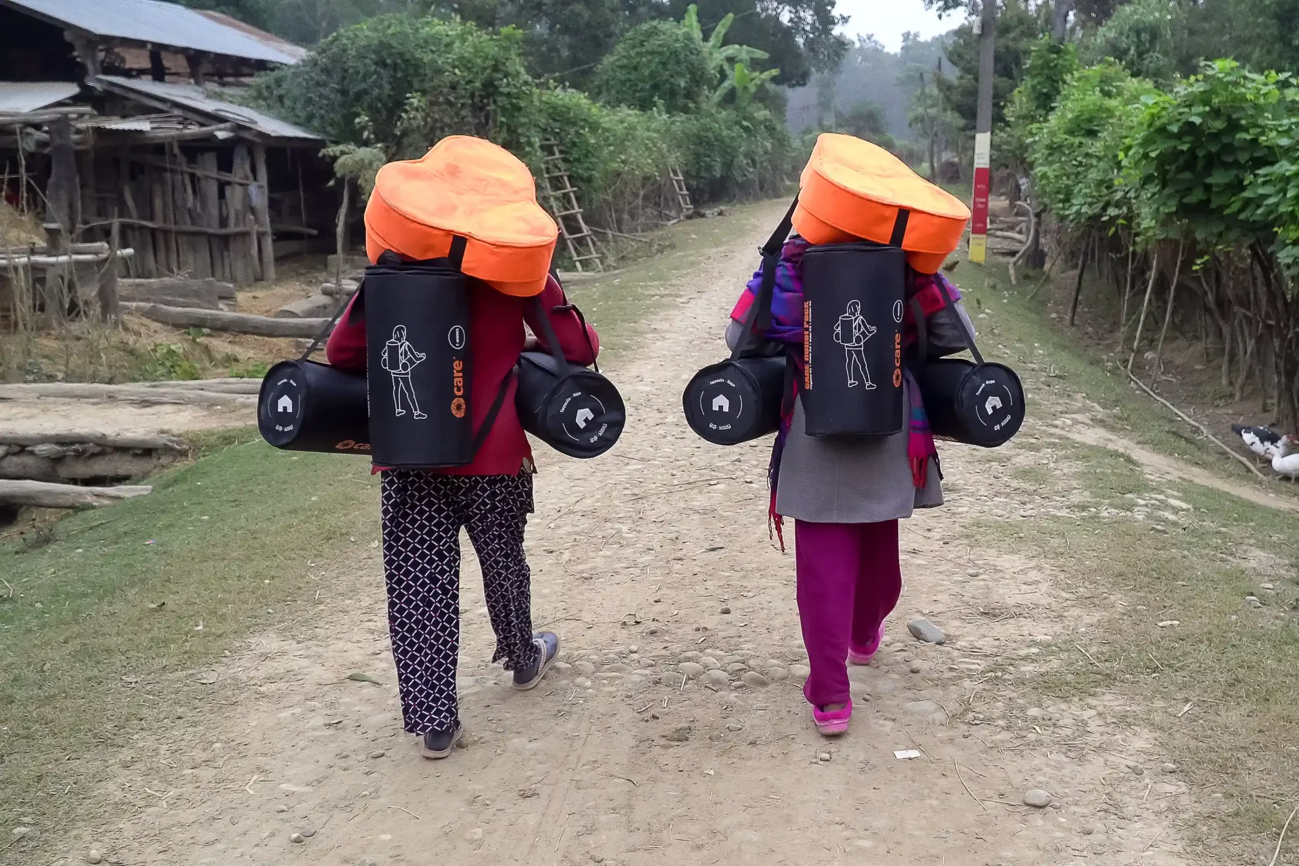 Two people in Nepal walk down a dirt path carrying CARE emergency backpacks with orange tops and black cylindrical bags.