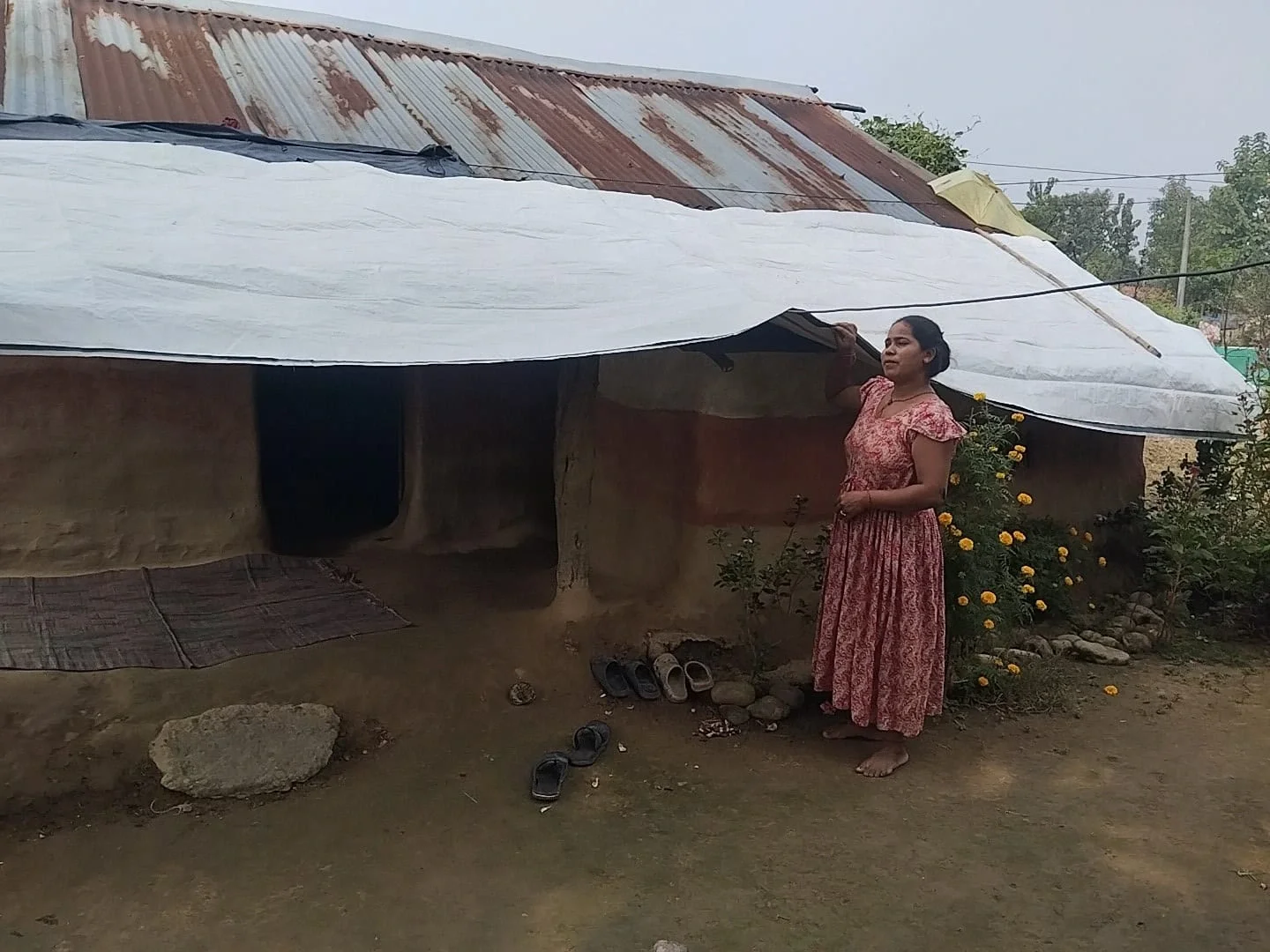 Alt text: A woman in Nepal stands outside a mud-walled shelter, adjusting a large white plastic tarp used to repair and cover a damaged corrugated metal roof.