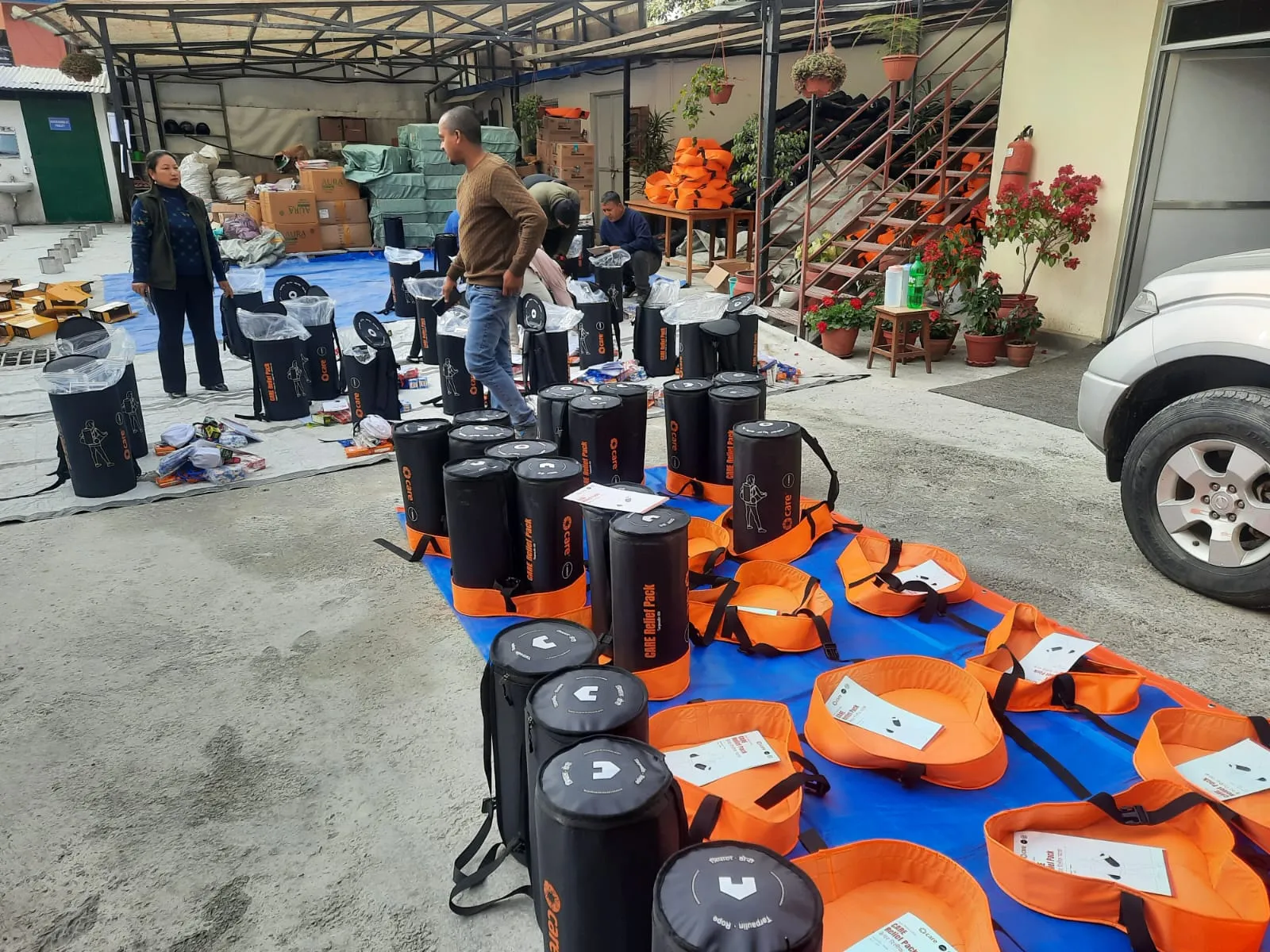 CARE workers assemble disaster relief kits, organizing rows of black CARE bags and orange backpacks on blue tarps.