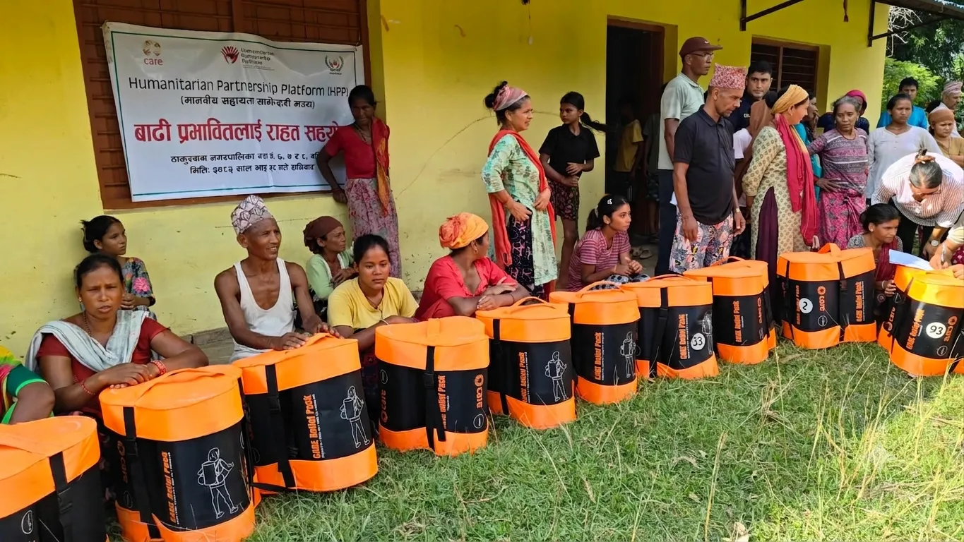 A group of people gathers outdoors in front of instructional posters and a banner reading Humanitarian Partnership Platform, with several orange and black CARE relief packs lined up on the ground.
