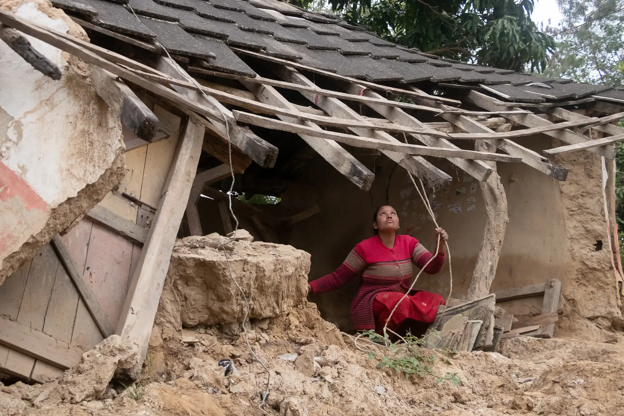 A Nepali woman in a striped sweater crouches inside a damaged mud house, reaching toward exposed rafters and broken roof tiles.
