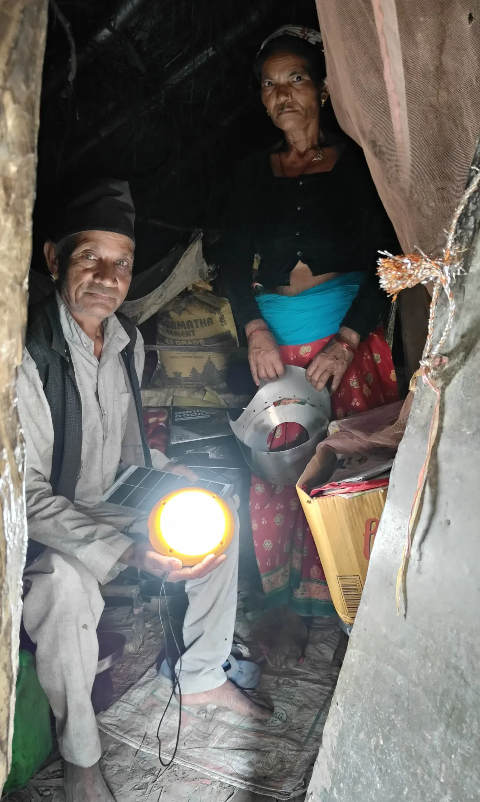 Inside a dim shelter, a man holds a glowing solar lamp connected to a small solar panel, while a woman stands behind him holding a metal stove component.