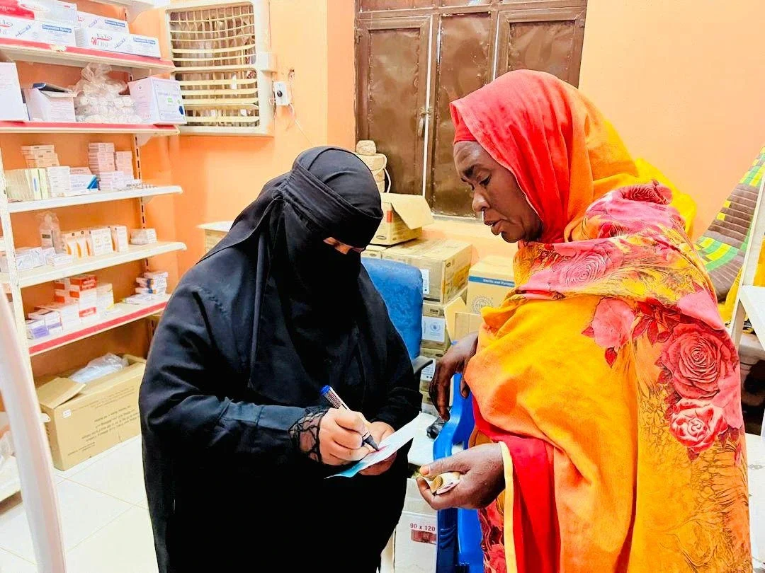 Inside a medical facility in Sudan, with shelves of medicine in the background, a healthcare worker wearing a black abaya and niqab writes on a document. A woman dressed in a vibrant orange and pink patterned headscarf and robe stands opposite her, watching attentively.