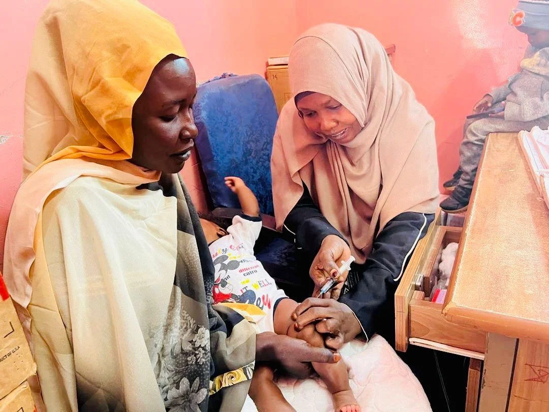 Inside a clinic in Khartoum, a healthcare worker wearing a light brown hijab carefully administers a vaccine injection into a young child's thigh.
