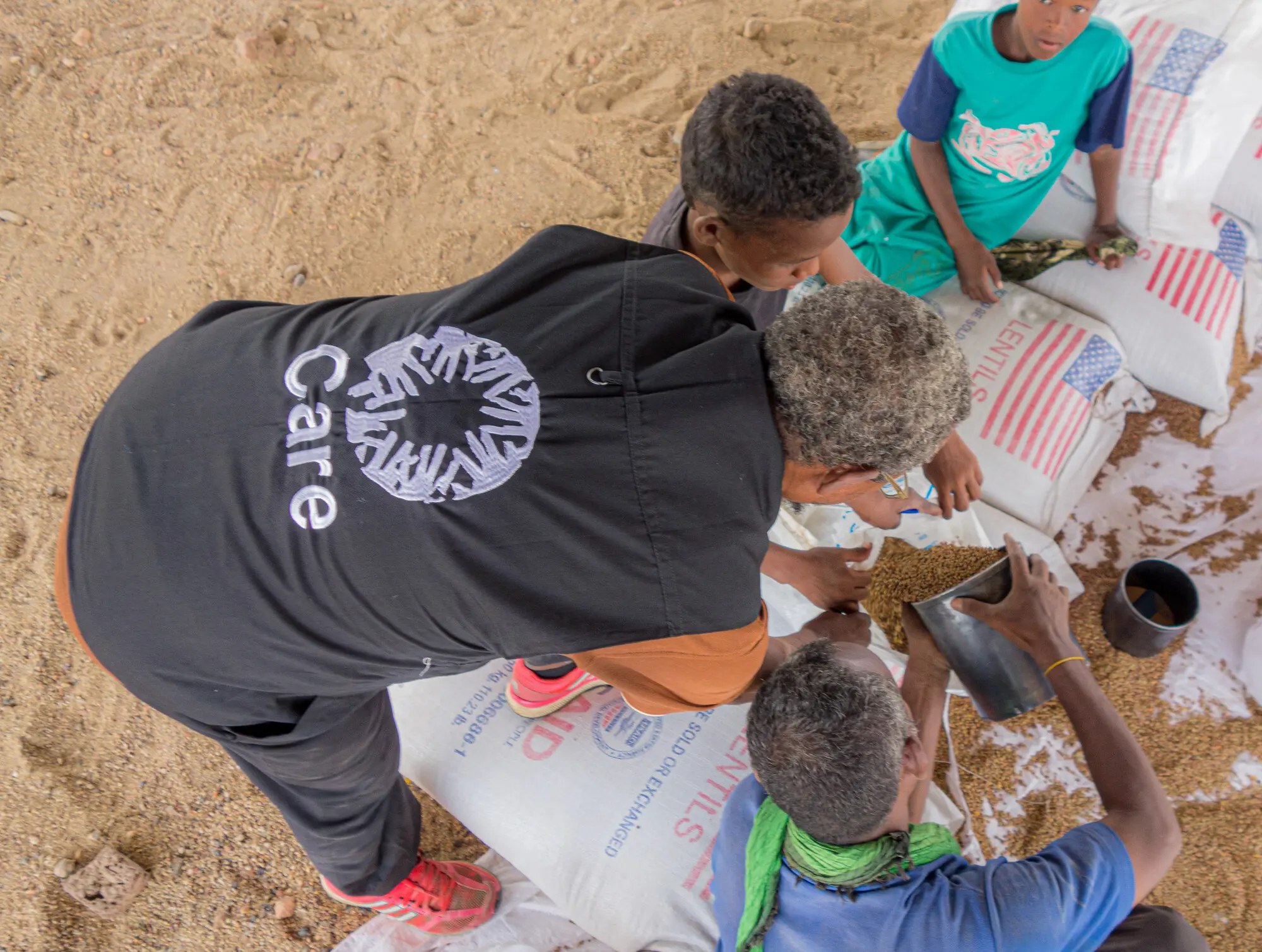 An overhead view shows a worker wearing a black CARE vest assisting people during a food distribution in Sudan. A person scoop lentils from a large white sack featuring an American flag design into a container, while children sit nearby on top of stacked supply sacks on the sandy ground.